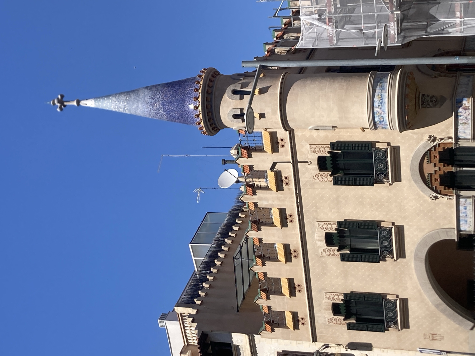 The image captures a close-up, low-angle view of the top section of a building in Barcelona, Spain, under a clear blue sky. The building features a distinctive turret with a conical roof that is covered in blue mosaic tiles, graduating in color from dark blue at the bottom to a lighter blue and white towards the pointed apex. At the very top of the turret is a four-pointed cross ornament. The turret is adorned with decorative ceramic tiles at its base and around its top rim.
Adjacent to the turret, the main facade of the building is visible, painted in a light beige or tan color. This section of the building has a patterned texture and is decorated with small, pointed crenellations along the top edge, interspersed with yellow and orange ceramic elements. Several windows with dark green shutters are visible, some with wrought-iron balconies. Below the windows, there are decorative arches and what appear to be additional ceramic tile details.
On the roof of the building, further to the left, there is a white balustrade and a dark metal railing. A modern glass structure is also visible on this part of the roof. A satellite dish and an antenna are mounted on the building. To the right of the turret, scaffolding covered with white netting is visible, with "OMEGA" printed on sections of the netting, suggesting ongoing construction or renovation work. A lamppost with a modern light fixture extends from the turret area, crossing in front of the scaffolding. The overall impression is of an architecturally interesting building in an urban setting.