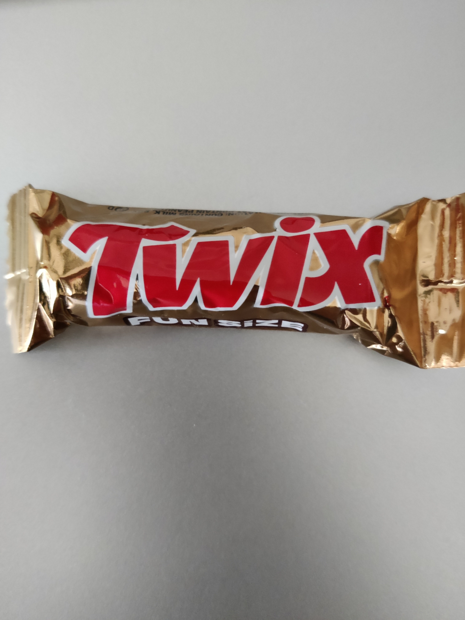 The image shows a single Twix candy bar in its golden wrapper. The Twix logo, in red and white, is prominently displayed on the front of the wrapper, along with the words "FUN SIZE" underneath. The wrapper is slightly crumpled, suggesting it has been handled. The candy bar is lying horizontally on a plain, light-colored surface, which appears to be a table or countertop. The background is out of focus and does not provide any contextual information about the environment. There are no people or other objects visible in the image. The lighting is even, casting no significant shadows. The image is a close-up shot of the candy bar. The location is the United States of America.