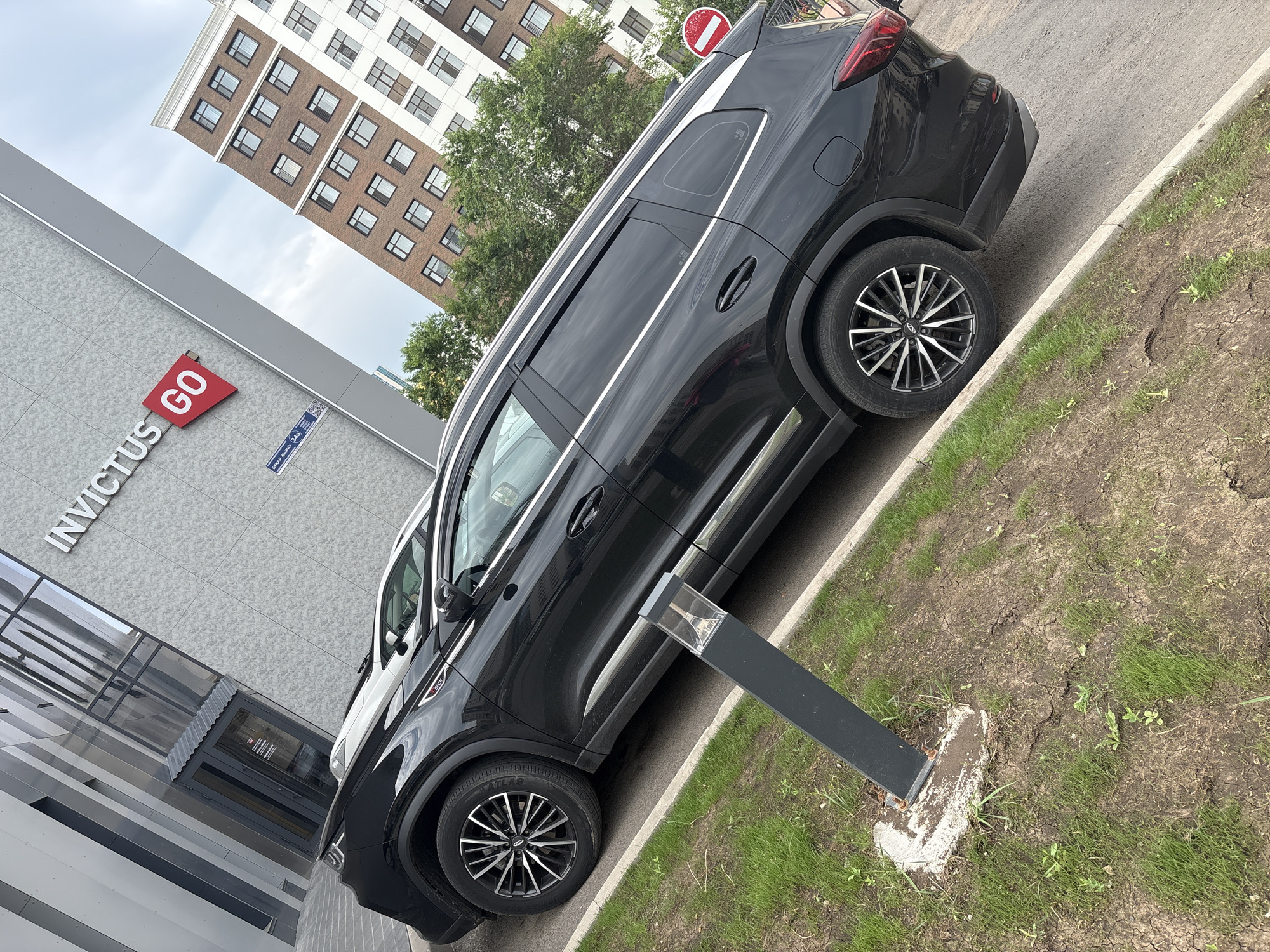 The image shows a black SUV parked on a street in Astana, Kazakhstan. The vehicle is a modern model with tinted windows and alloy wheels. In the background, there is a modern building with the text "INVICTUS GO" on its facade. Another residential building with many windows is also visible, along with some trees. The sky is overcast. A red and white circular sign is visible to the right of the car. The street appears to be made of asphalt, and there is a strip of grass next to it, with some cracked earth and small plants. A modern-looking lamppost is planted in the grass. The scene is captured from a slightly elevated angle, looking down towards the car and the street.