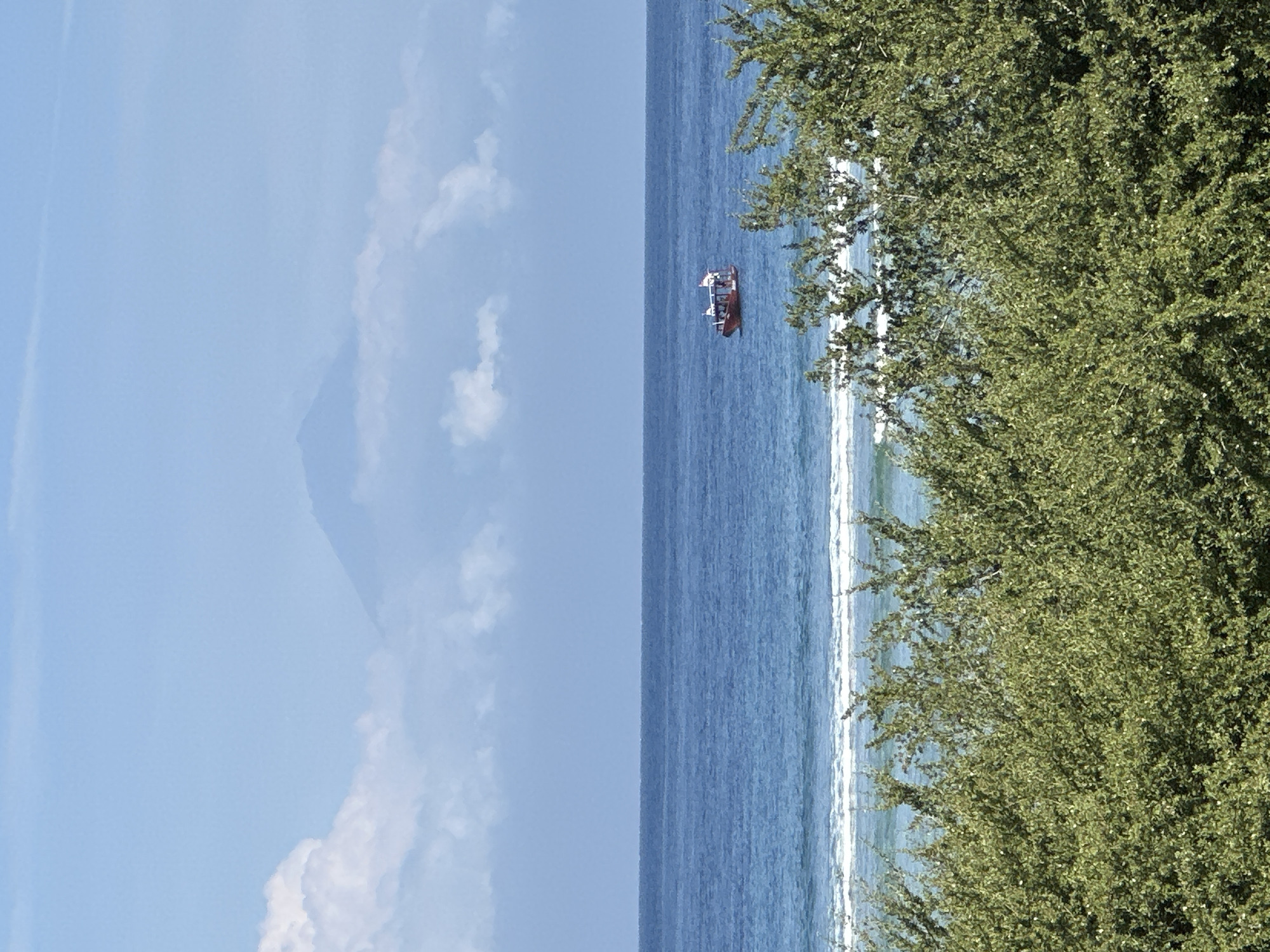 The image depicts a coastal scene in Unknown, Indonesia. A small boat is in the sea. To the right, green trees and bushes suggest a shoreline. The sky is blue with scattered clouds. A mountain or large hill is partially visible on the left horizon.