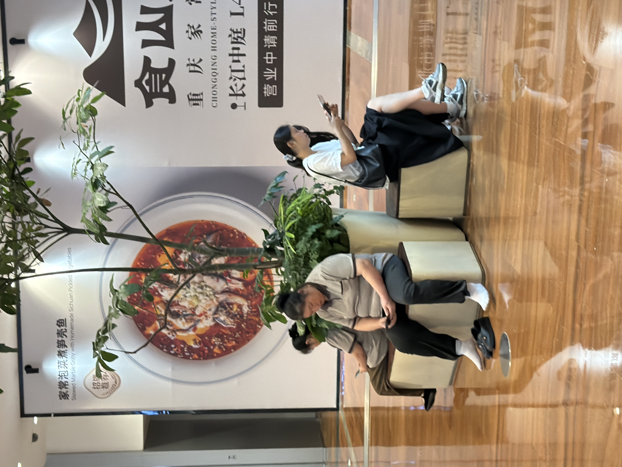 In the Yuzhong District, China, three individuals are seated on benches in a commercial space. The environment features a polished wooden floor reflecting the overhead lighting and a large white banner with Chinese text and imagery of food. A potted plant is positioned between two of the individuals.
On the left, two individuals are seated side-by-side. The person on the far left is facing away from the camera, looking down at a mobile device held in their hands. They are wearing a grey t-shirt and brown pants, with black shoes. Next to them, the second person is seated facing forward, also looking down at a mobile device. This individual has their hair pulled back in a bun and is wearing a grey polo shirt and black pants, with white socks and black slip-on shoes.
On the right, a third individual is seated facing forward, holding a mobile device and looking at it. They are wearing a white t-shirt and a black skirt, with white socks and athletic shoes. A black handbag is slung over their shoulder.
The banner behind them has prominent Chinese characters and an image of a bowl of food. The text on the banner appears to advertise a restaurant or food establishment. The lighting in the space is bright, creating reflections on the glossy floor.