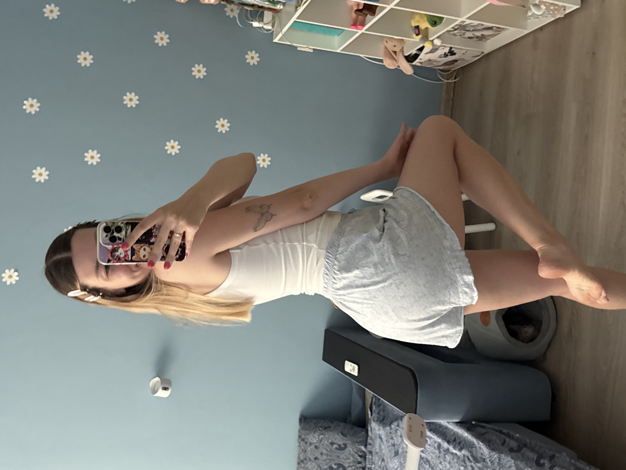 A young woman with blonde hair, wearing a white tank top and grey shorts, takes a selfie in a room with light blue walls decorated with daisy stickers. She is holding a phone with a cat-themed case and has a butterfly tattoo on her left arm. In the background, a bed with a patterned comforter is visible, along with a dark grey object, a cat bed with a cat inside, and a white shelving unit filled with toys. The floor is light wood laminate. The scene appears to be an indoor residential setting, likely a bedroom. There is no indication of the specific location within Moscow, Russia.
