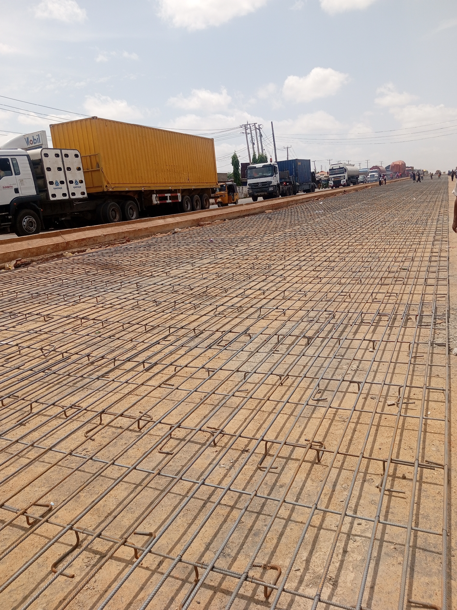The image depicts a road construction site in an unidentified urban location. The foreground is dominated by a grid of steel rebar laid out on the ground, prepared for concrete pouring. Beyond the rebar, a line of vehicles, including container trucks and other heavy-duty vehicles, are visible, suggesting traffic congestion or a temporary stop due to the road work. In the background, utility poles and power lines are visible against a partly cloudy sky. Several people are standing or walking near the edge of the construction zone. The general environment suggests an urban or suburban setting undergoing infrastructural development.