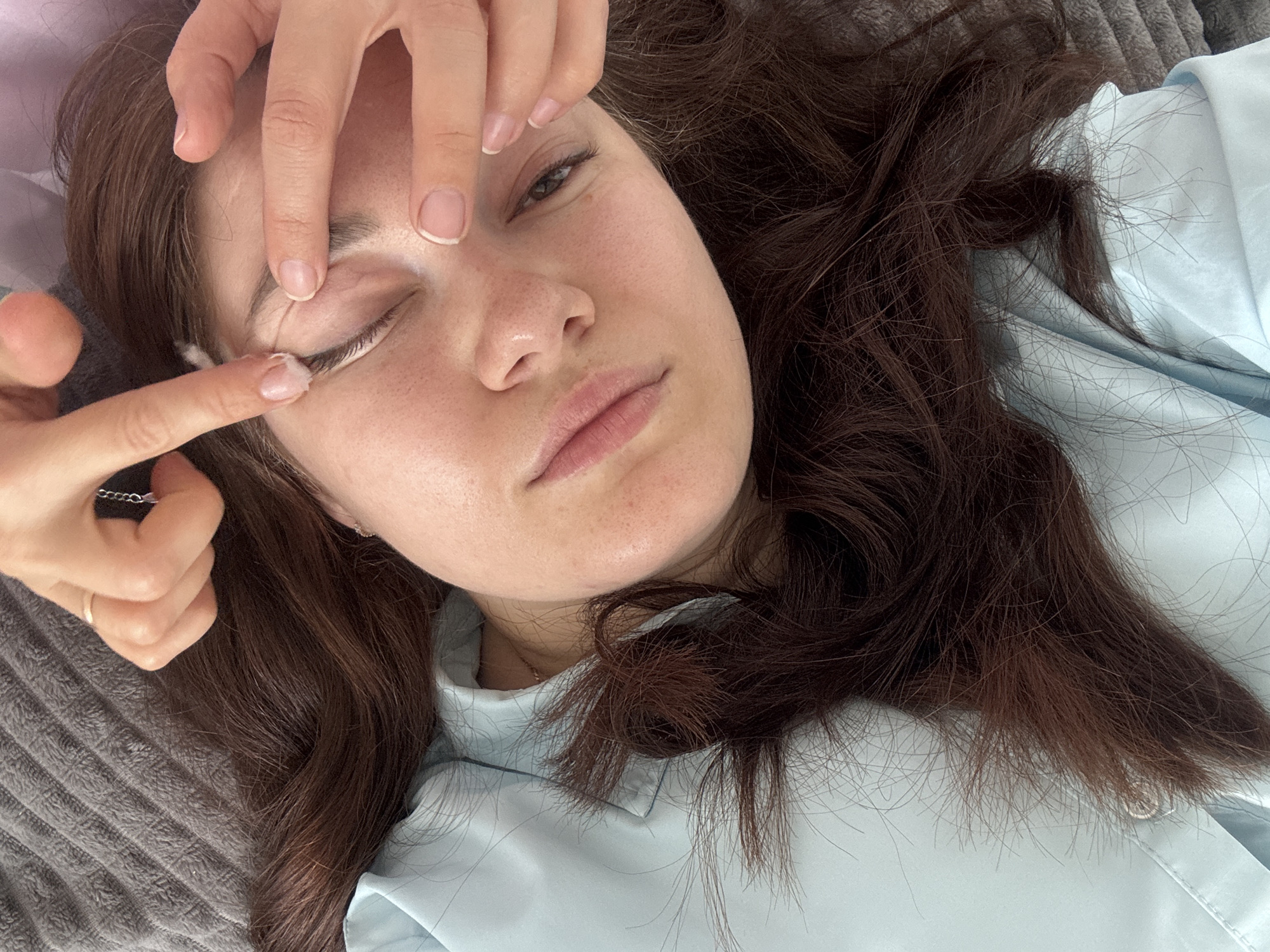 A young woman with long, dark brown hair is lying down with her head on a grey, textured surface, possibly a bed or couch. She is wearing a light blue pajama top. Her eyes are partially closed as someone's hands are gently applying cream or product to her eyebrow area. Her facial expression is relaxed. The environment appears to be indoors. There is no indication of the city of Chaikovsky, Russia, in the image.