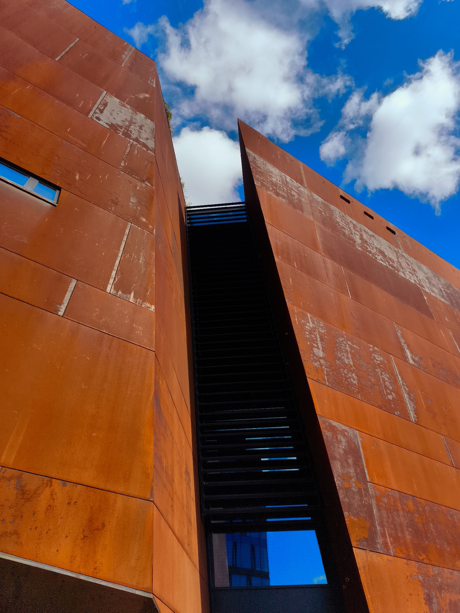 The image depicts a low-angle shot of a modern building with large, rust-colored metal panels. The building has a sharp, angular design, with two facades meeting to create a deep recess. The recess reveals a dark, vertical structure with horizontal slats, suggesting a staircase or ventilation system. A small, rectangular window with a blue tint is visible on the left facade. The sky above is a vibrant blue with scattered white clouds. The architecture is industrial and striking. This building is the European Solidarity Centre in Gdańsk, Poland, a museum and library commemorating the Solidarity movement.