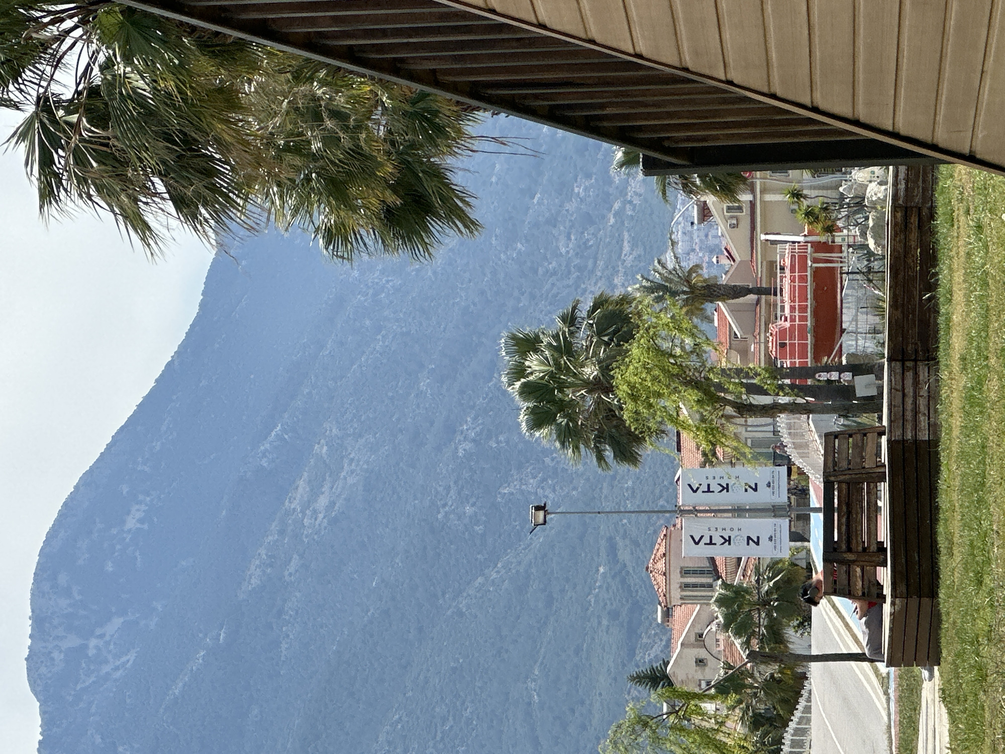 The scene is a view from under a covered structure, possibly a shelter or veranda, in Fethiye, Turkey. A large, forested mountain dominates the left side of the frame. In the center and right, the scene shifts to a residential area with buildings, palm trees, and street signage that reads "NOKTA HOMES". A road runs between the buildings and a grassy area. A wooden bench is visible near the edge of the covered structure. An individual is sitting on the bench.