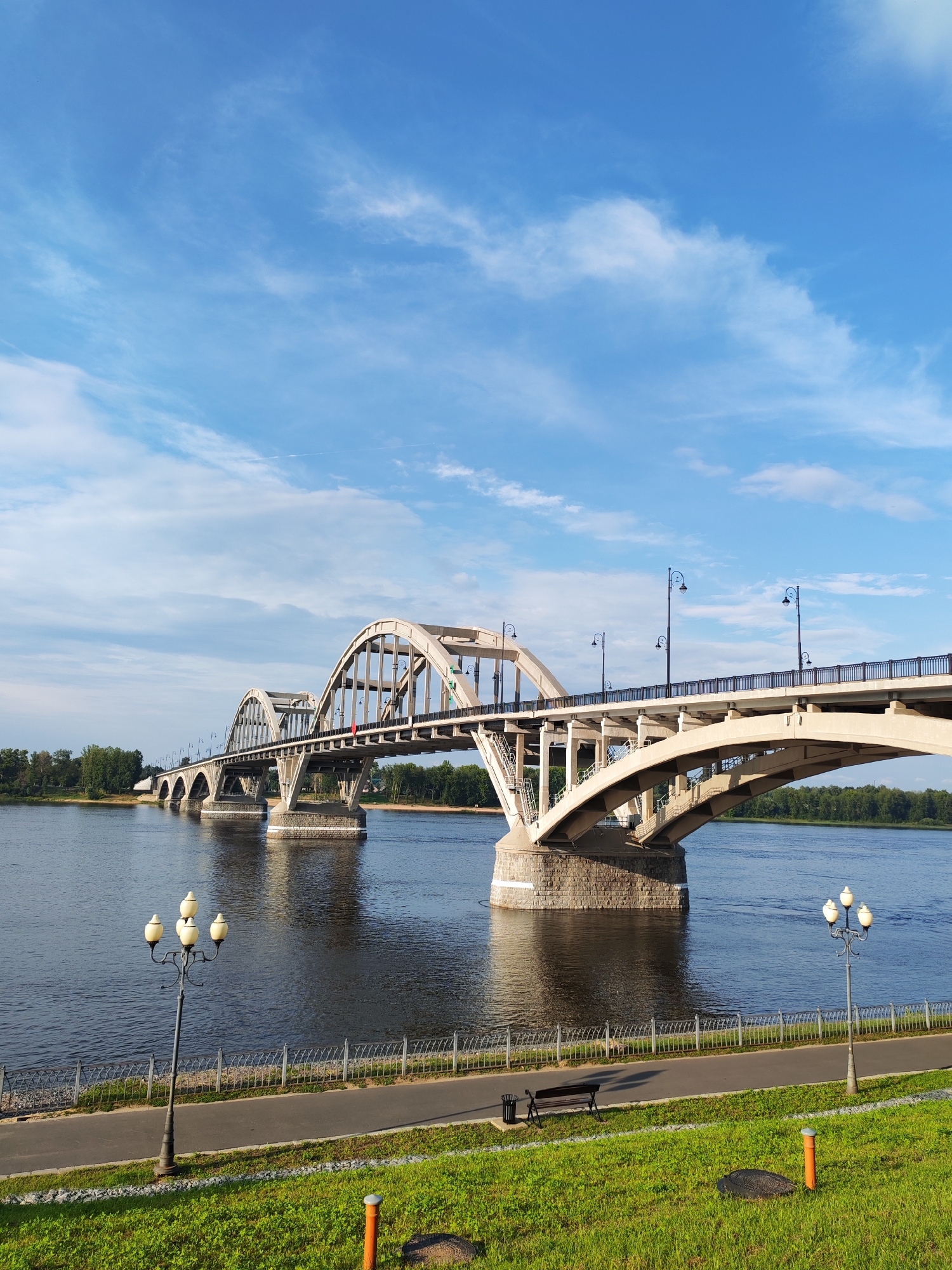 The image depicts a wide concrete arch bridge spanning a body of water, likely a river, under a clear blue sky with scattered wispy clouds. The bridge features multiple arches and is supported by robust stone pillars. Along the near bank of the river, there is a paved pathway, a metal fence, a park bench, a trash receptacle, and lampposts. Lush green grass covers the foreground. In the distance, dense green trees line the opposite bank of the river, and the bridge extends far into the background, suggesting a significant structure. The scene appears to be in a park or waterfront area of the city of Bol'shie Mkhi, Russia.