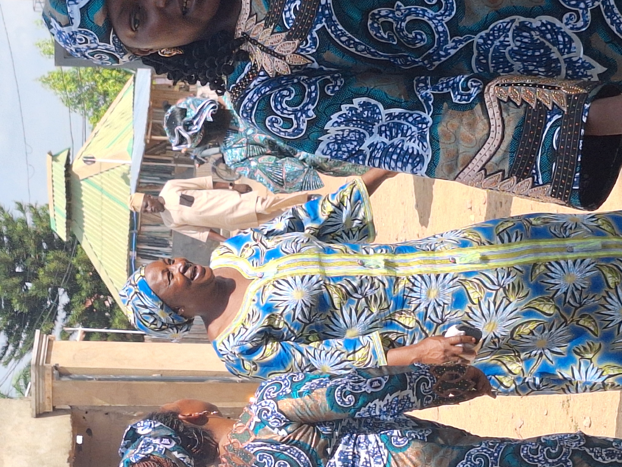 In Sabon Tasha, Nigeria, the image captures a group of people in an outdoor setting, likely a street. The focus is on several women dressed in traditional African attire with vibrant blue and floral patterns. One woman is gesturing and seems to be speaking, while others are visible in the background. A man in a light brown uniform is also present in the background, partially obscured. The environment appears to be a somewhat dusty or sandy area, with buildings and foliage visible in the background.