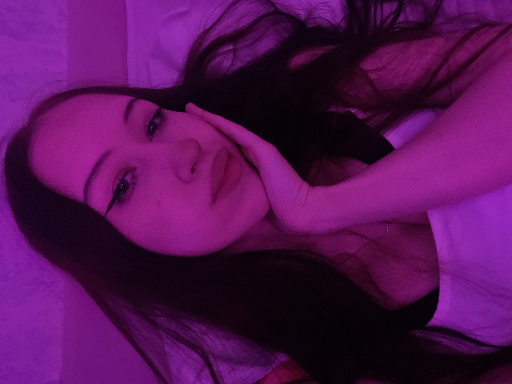 A young woman with long, dark hair is resting on her side, looking towards the camera. She is wearing a white top. Her right hand is resting on her cheek, and she has winged eyeliner. The lighting in the image is a vibrant pink, casting a strong glow on her skin and surroundings. The background appears to be a textured surface, possibly a wall, also illuminated by the pink light. There is a hint of a white sheet or bedding behind her. The image is a close-up, focusing on the woman's face and upper body. The specific location in Votkinsk, Russia, cannot be determined from the visual information provided.