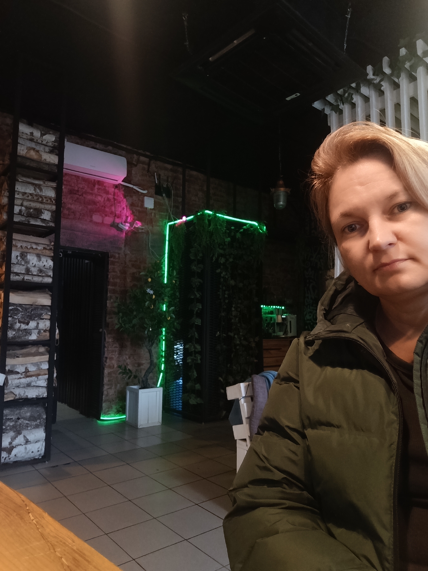 A woman with short, blonde hair and a serious expression is in the foreground, wearing a green puffer jacket. She appears to be taking a selfie in a restaurant or cafe.
The background is dimly lit with a mix of industrial and natural elements. A brick wall is visible with a pink neon light above a doorway. To the right, green neon lights highlight a vertical plant display. On the left, a metal-framed shelf is filled with stacked birch logs. The floor is tiled. A potted lemon tree sits near the green neon display. In the mid-ground, are objects that look like an espresso machine and coffee mugs.
Based on the information provided, the location is Chaikovsky, Russia.