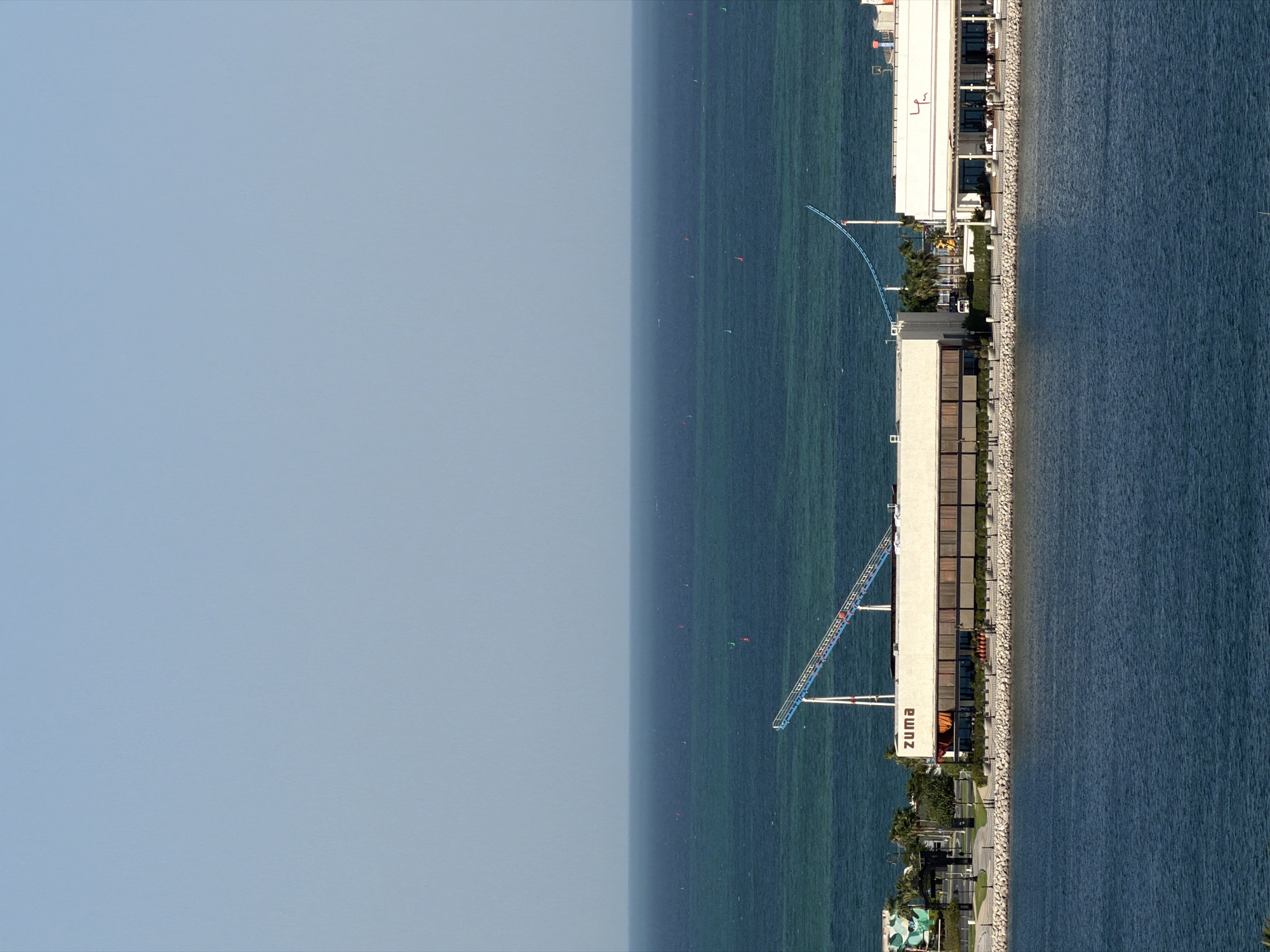 This is a vertical, eye-level aerial shot of a modern building complex situated on the edge of a large body of water. The water is a deep blue and appears calm, with a faint green hue in some areas, suggesting it is relatively shallow. The building is long and rectangular with a white facade and numerous dark-tinted windows. The word "zuma" is visible in large, vertical, white lettering on the side of the building.
A pathway paved with light-colored stones runs along the water's edge, separating the building from the water. There are some trees and landscaping visible around the building, including palm trees. Further out in the water, a series of small, colorful buoys (red and green) are scattered, possibly marking a course or an area.
A prominent feature is a blue and white structure extending from the building over the water, resembling a slide or a bridge. The sky is a pale, hazy blue, suggesting either early morning or late afternoon, or possibly a slightly overcast or foggy condition. The overall scene conveys a sense of leisure and a well-developed waterfront area. There are no visible people in the image.