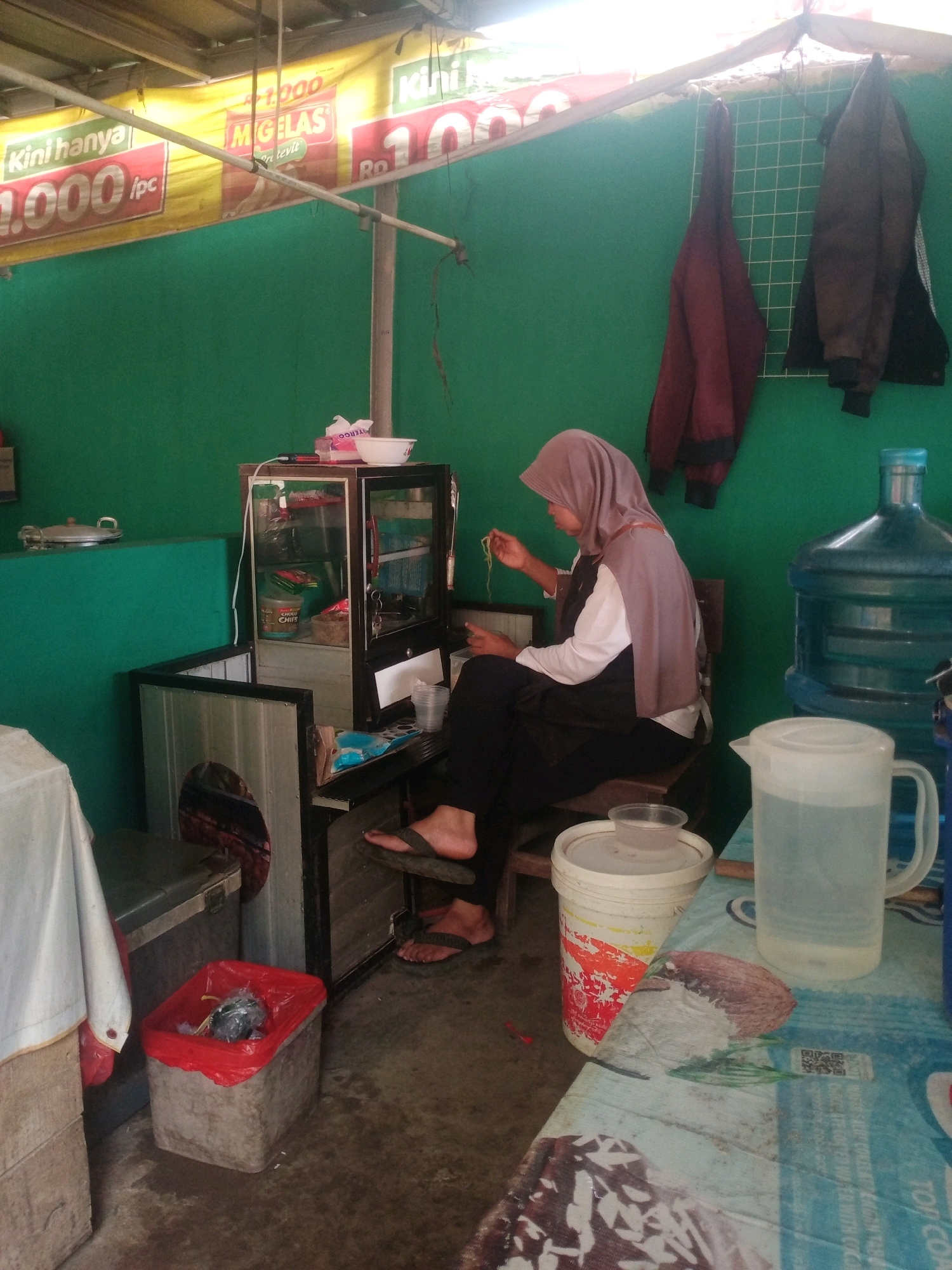 The image depicts an outdoor food stall, likely in Bandar Lampung, Indonesia, as suggested by the yellow banner with Indonesian text advertising prices like "1.000 Rp" and "10.000 Rp". A woman wearing a light brown hijab and a white shirt is seated on a wooden stool, tending to food. She is holding a strand of yellow noodles in her right hand, appearing to be preparing or eating them. In front of her is a small glass display case containing various food items and containers, some labeled with Indonesian text. To her right, a large blue water cooler jug and a white pitcher are visible on a table covered with a blue and white patterned tablecloth. Behind the woman, two jackets are hanging on a grid. The stall has a green wall as a backdrop. To the left, a trash bin with a red liner is present, and a white tablecloth covers a surface. The overall impression is a humble, operational food vending stall.