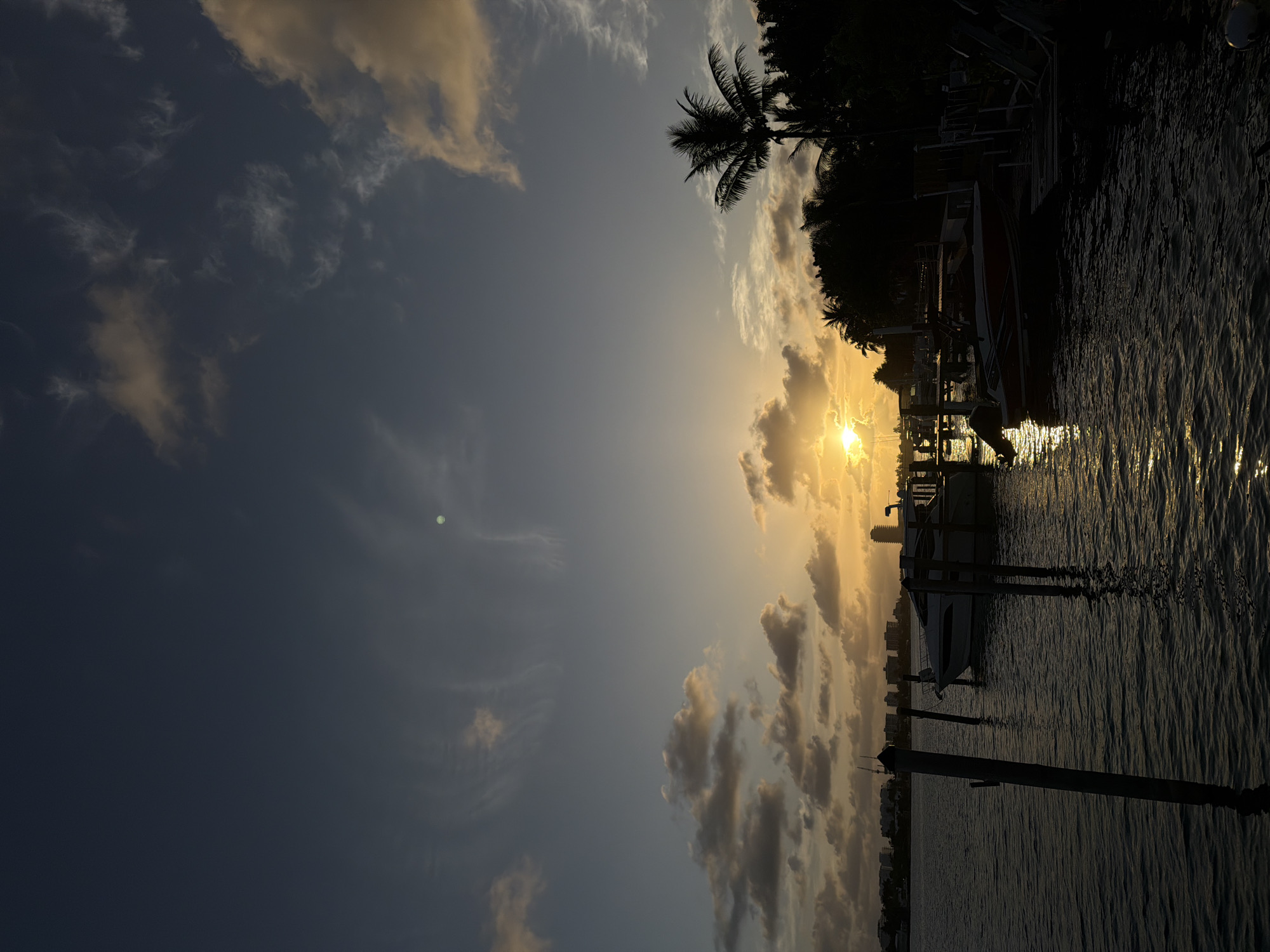 The photograph captures a late afternoon or early evening scene, likely a sunset, in Miami Beach, United States of America. The orientation of the image is rotated counterclockwise. The sun's radiant glow is positioned toward the center, partially obscured by clouds, with a palm tree and the silhouettes of buildings and other vegetation visible in the background. In the foreground, there's a body of water, possibly a bay or waterway, reflecting the sun's light in a shimmering pattern. Several boats are docked along a pier or a shoreline. Wooden posts stand prominently in the water near the bottom of the frame, adding to the waterfront ambience. The sky is a gradient of colors, from lighter hues near the sun to darker shades towards the top left, suggesting the transition of daylight into dusk.