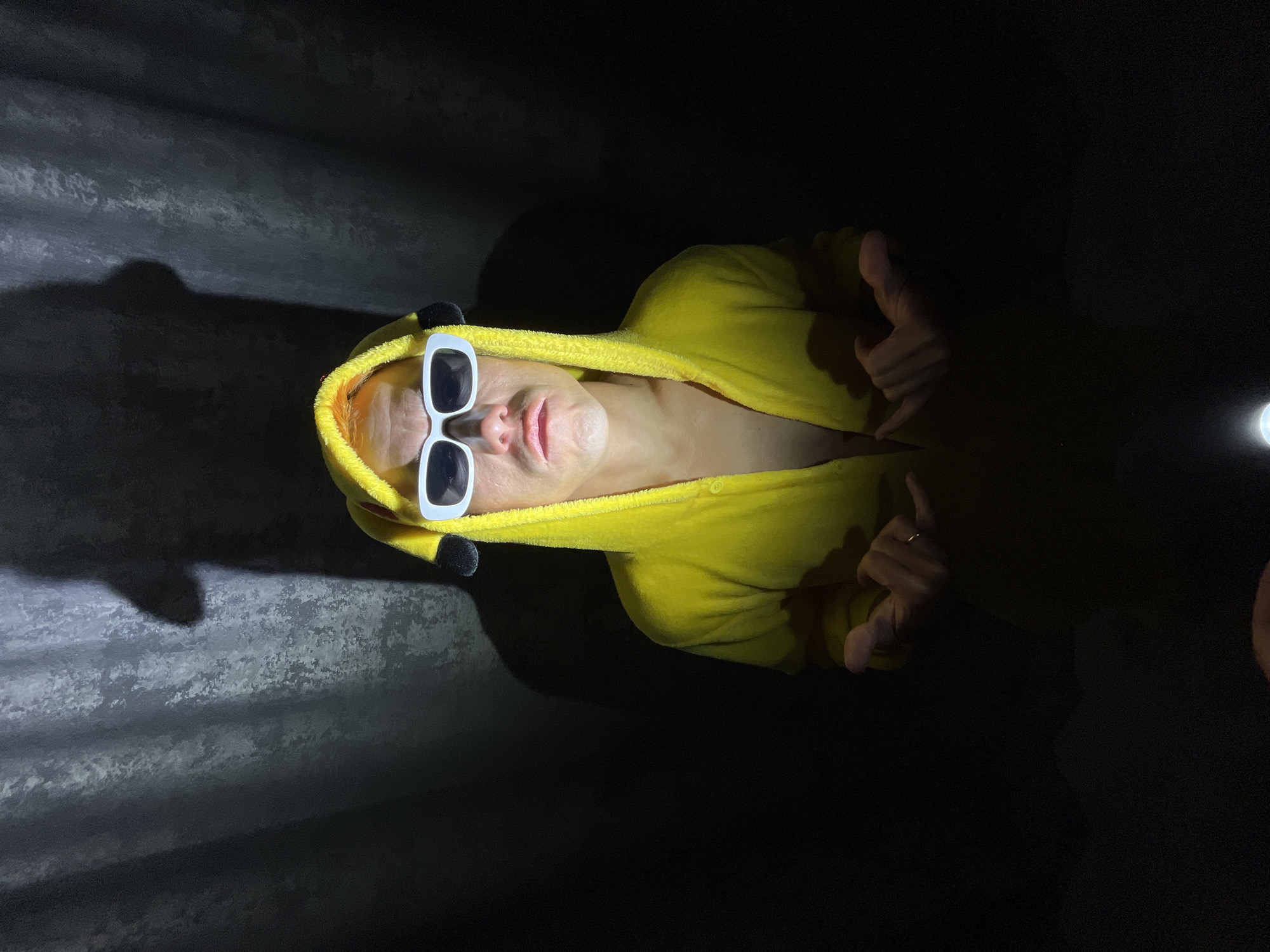 A person wearing a yellow Pikachu onesie with white sunglasses is lying down. The photo is taken in dim lighting, casting shadows on the corrugated metal surface on the left side of the frame. The person is giving a "shaka" sign (hang loose) with both hands. A light source on the right side illuminates the person slightly. No information in the image to confirm the location in the city of Chaikovsky, Russia.