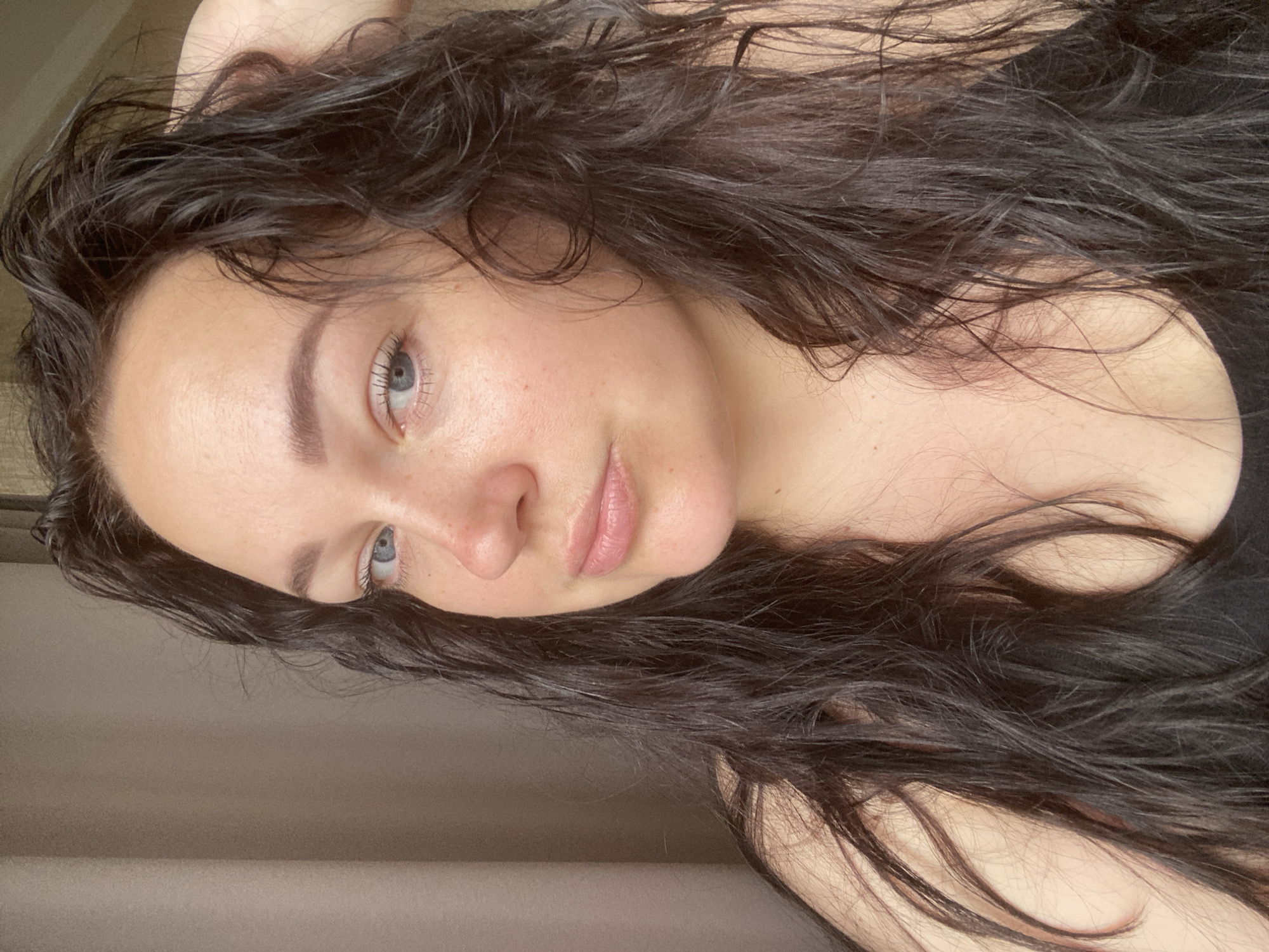 A medium, angled, indoor shot depicts a woman with long, dark, wavy hair. She has fair skin, light blue eyes, and full lips. Her expression is neutral. The background is muted, with a beige wall and a hint of what appears to be a dark top. The location cannot be determined as Sochi, Russia, as there are no elements indicating the location.