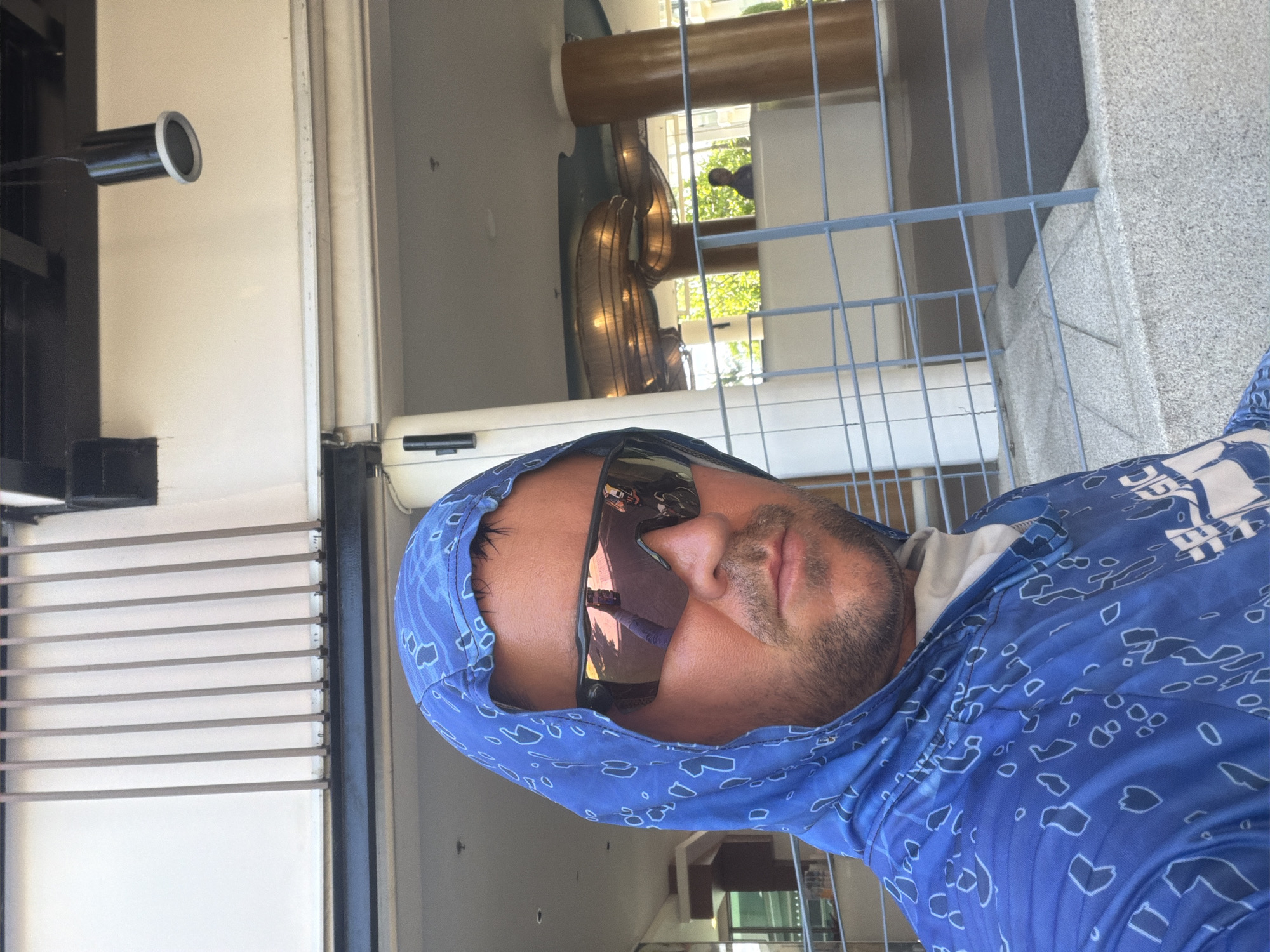 In Baan Chaweng, Thailand, a man wearing a blue hooded shirt with a white pattern and sunglasses is taking a selfie. He has facial hair and is looking directly at the camera. Behind him, a building with vertical white louvers and dark horizontal beams is visible. A cylindrical black light fixture hangs from the ceiling. Further back, a reception desk with a decorative curved element and wooden columns can be seen. In the background, a person is standing near trees. The foreground shows a stone staircase with a grey metal railing and a dark mat.