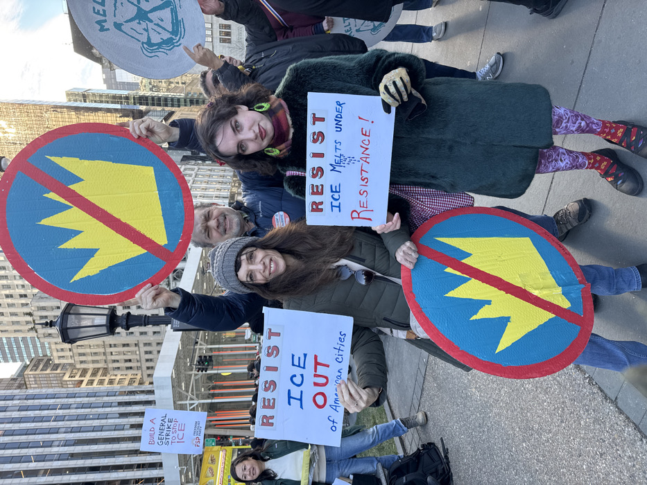 A group of people are protesting. They hold signs and circular placards. The signs read "RESIST ICE OUT of American cities", "RESIST ICE MELTS UNDER RESISTANCE!", and "BUILD A GENERAL STRIKE TO STOP ICE." The circular placards have a yellow arrow-like shape, crossed out with a red line, over a blue background with a red border. The backdrop includes buildings and other protesters. One protester is wearing red and purple patterned leggings.