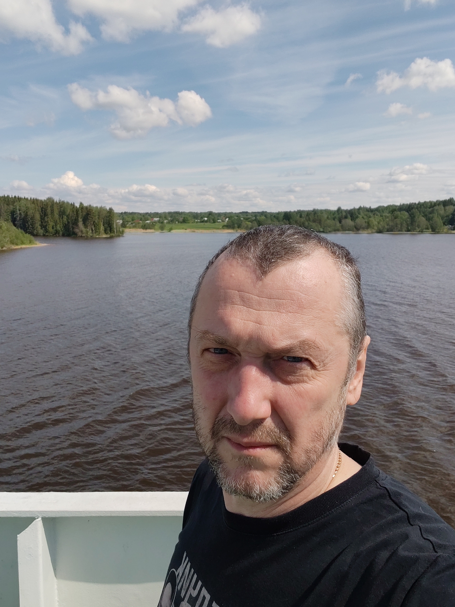 A middle-aged Caucasian man with short, graying hair and a trimmed beard stands on what appears to be the deck of a boat or ferry. He is wearing a black t-shirt and a gold chain is visible around his neck. The background reveals a body of water, likely a lake or wide river, bordered by forested areas. The sky is partly cloudy with patches of blue. Based on the prompt, the location is Kondratovo, Russia. The man is facing the camera, suggesting he is taking a selfie. The presence of a railing suggests the person is standing on a boat and the surrounding bodies of water are indicative of an outdoor setting.