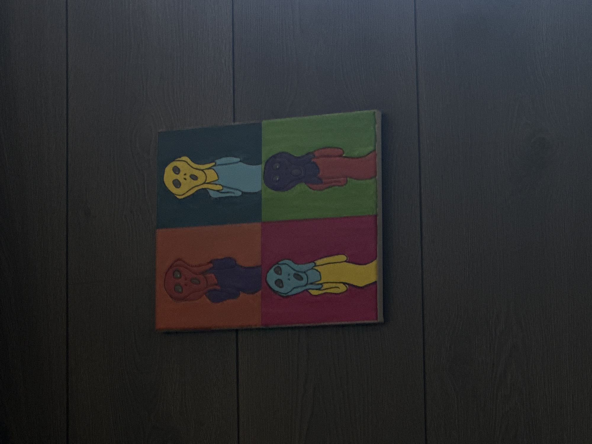 A four-paneled art piece hangs on a dark wood-paneled wall. The art piece features a depiction of an alien-like head in each panel, each colored differently against contrasting backgrounds. The top left panel shows a yellow head against a teal background. The top right panel shows a purple head against a green background. The bottom left panel shows a red head against an orange background. The bottom right panel shows a teal head against a pink background. The scene is dimly lit. There is no indication the photograph was taken in Huixquilucan de Degollado, Mexico.