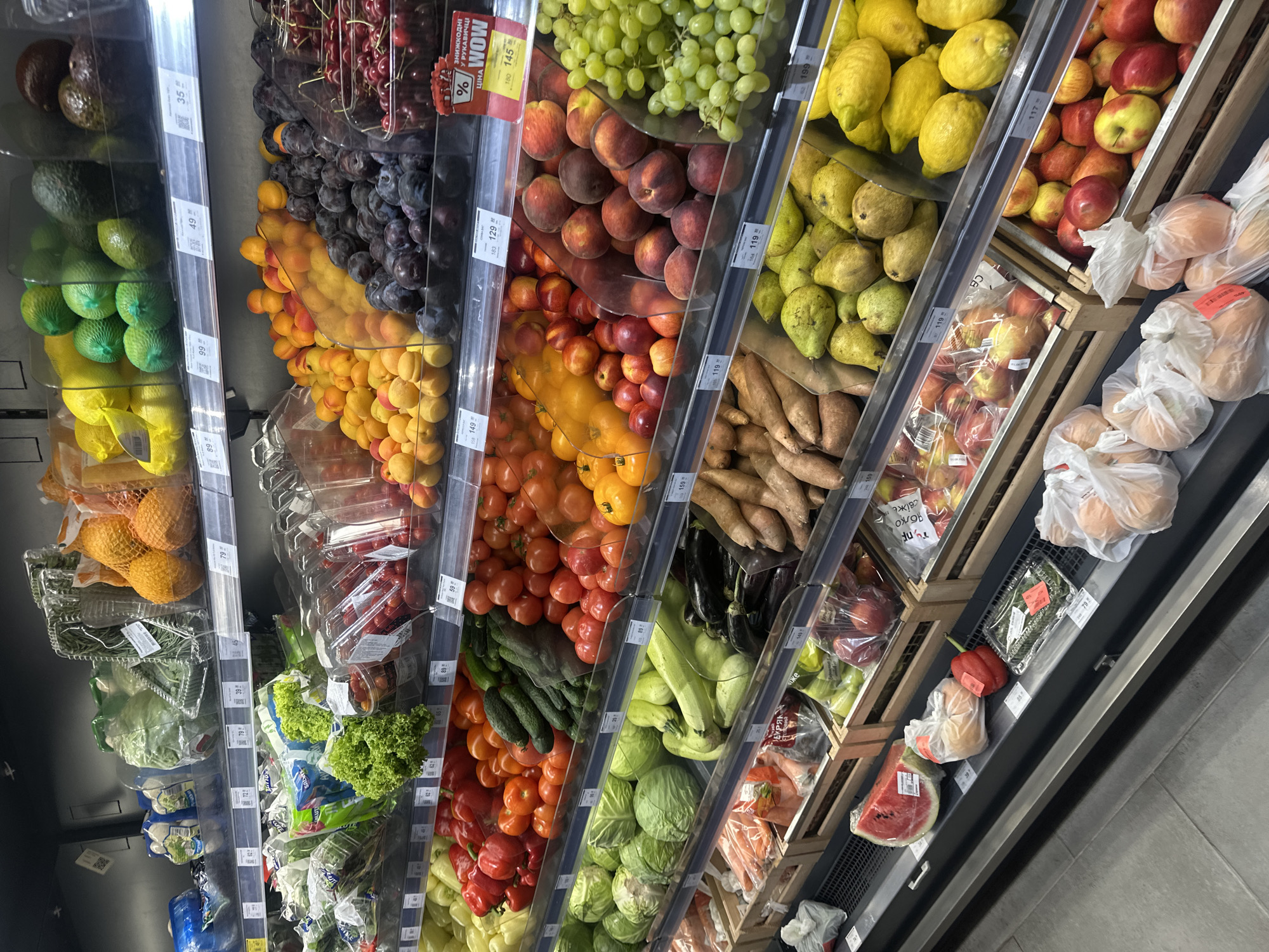 The image shows a close-up, eye-level shot of produce displayed on shelves in a supermarket in Ternopil, Ukraine. The lighting is bright and artificial, typical of a retail environment, illuminating a wide variety of fruits and vegetables.
On the top shelf, to the left, are packages of leafy greens, including what appears to be iceberg lettuce and possibly cauliflower. Further to the right, there are oranges and lemons in netting, and then several varieties of avocados and green, ball-shaped fruits which could be limes or small melons.
The middle shelf is laden with more produce. On the left, there are bags of salad greens and lettuce. Moving right, there are rows of red bell peppers, tomatoes, cucumbers, and other types of tomatoes. Further to the right, there are bins filled with plums, apricots, nectarines, peaches, and green grapes. There are also containers of red cherries.
The lower shelves display a variety of vegetables and fruits. On the left, there are cabbages, zucchini, and eggplants. In the center and right, there are sweet potatoes, pears, lemons, and apples. In the foreground, a large slice of watermelon sits on the bottom display shelf, accompanied by bags of onions and a red bell pepper.
Price tags are visible on several shelves, indicating prices in local currency. The overall impression is a well-stocked grocery store aisle. There are no people visible in the image. The scene is focused on the products themselves, with the shelves and lighting forming the environment.