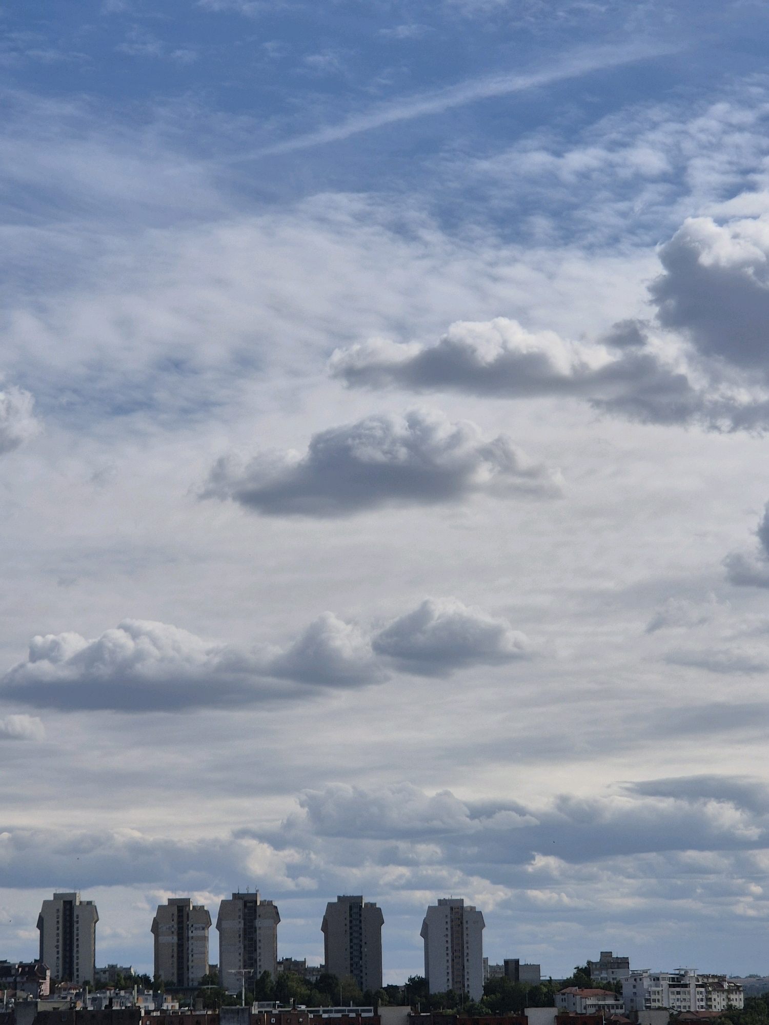 In the city of Unknown, Unknown, a panoramic view captures a sky filled with a mix of white and grey clouds against a light blue backdrop. A thin, wispy cloud formation stretches diagonally across the upper portion of the frame. Below the sky, a line of uniformly built, tall apartment buildings stretches horizontally across the mid-ground. These buildings are light-colored and feature numerous windows. In the foreground, a lower-rise cityscape with various residential buildings and greenery is visible. The overall scene suggests an urban environment under a cloudy sky.