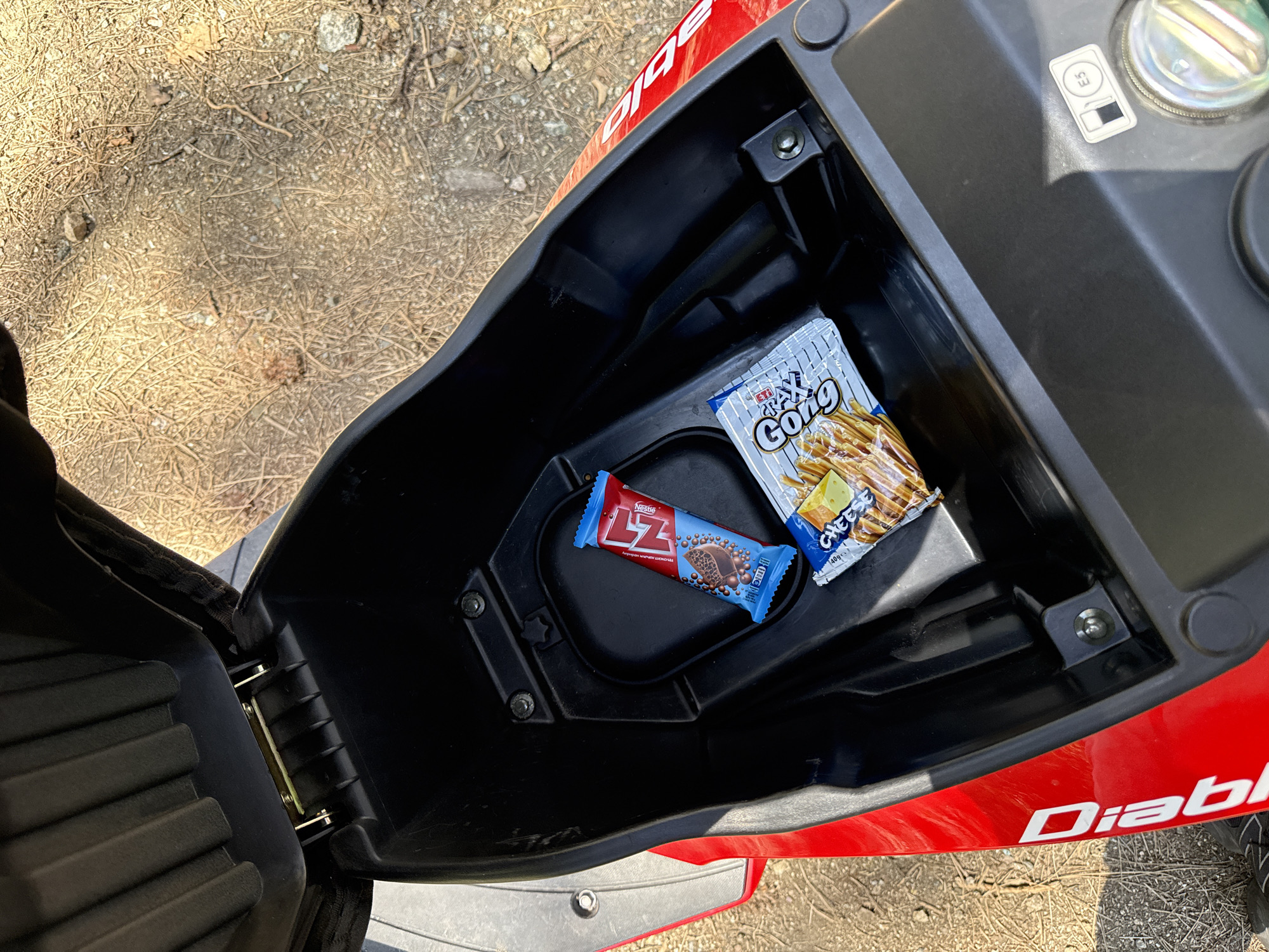 The image shows the opened storage compartment of a red "Diablo" scooter. Inside the compartment are two items: a "Nestle Lz" chocolate bar in a blue wrapper and a package of "Gong" cheese-flavored snack sticks. The compartment is black plastic, and there are metal bolts visible securing it to the scooter frame. The scooter is parked on a surface covered in dry pine needles and other debris. The image provides no information about Boboshevo or any human activity.