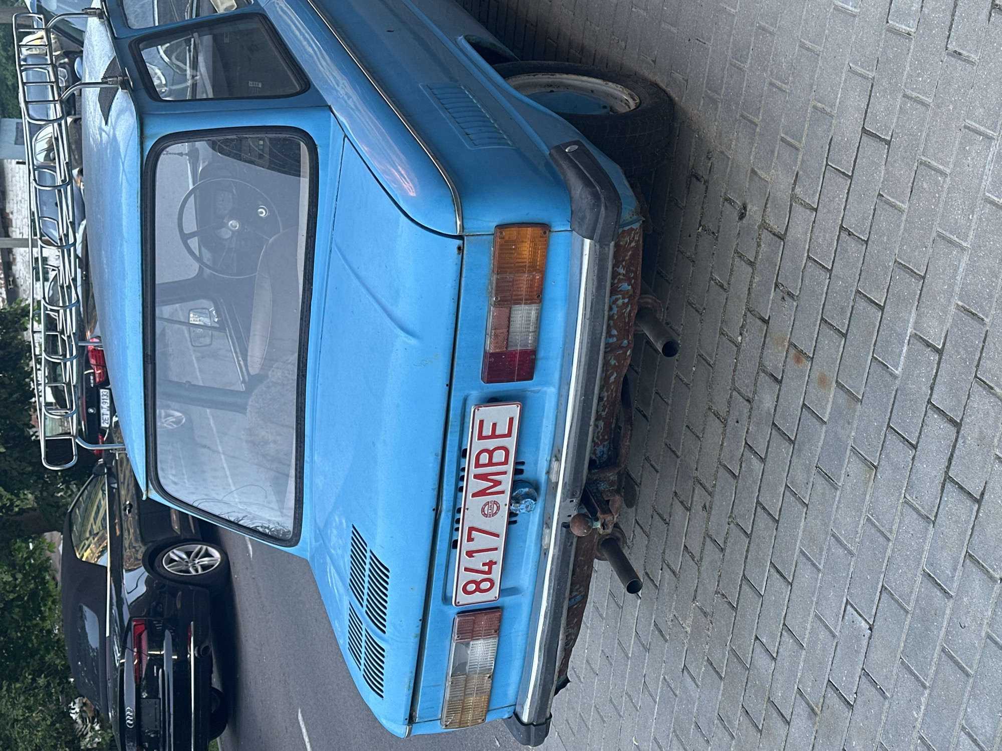The image shows the rear of a light blue vintage car parked on a paved surface. The car has a luggage rack on the roof. A license plate is visible, reading "8417 MBE" with a Minsk, Belarus seal. The car has visible signs of age and rust. Behind the blue car, a black car is parked on the asphalt. The scene is outdoors, likely in Minsk, Belarus, as indicated by the license plate.