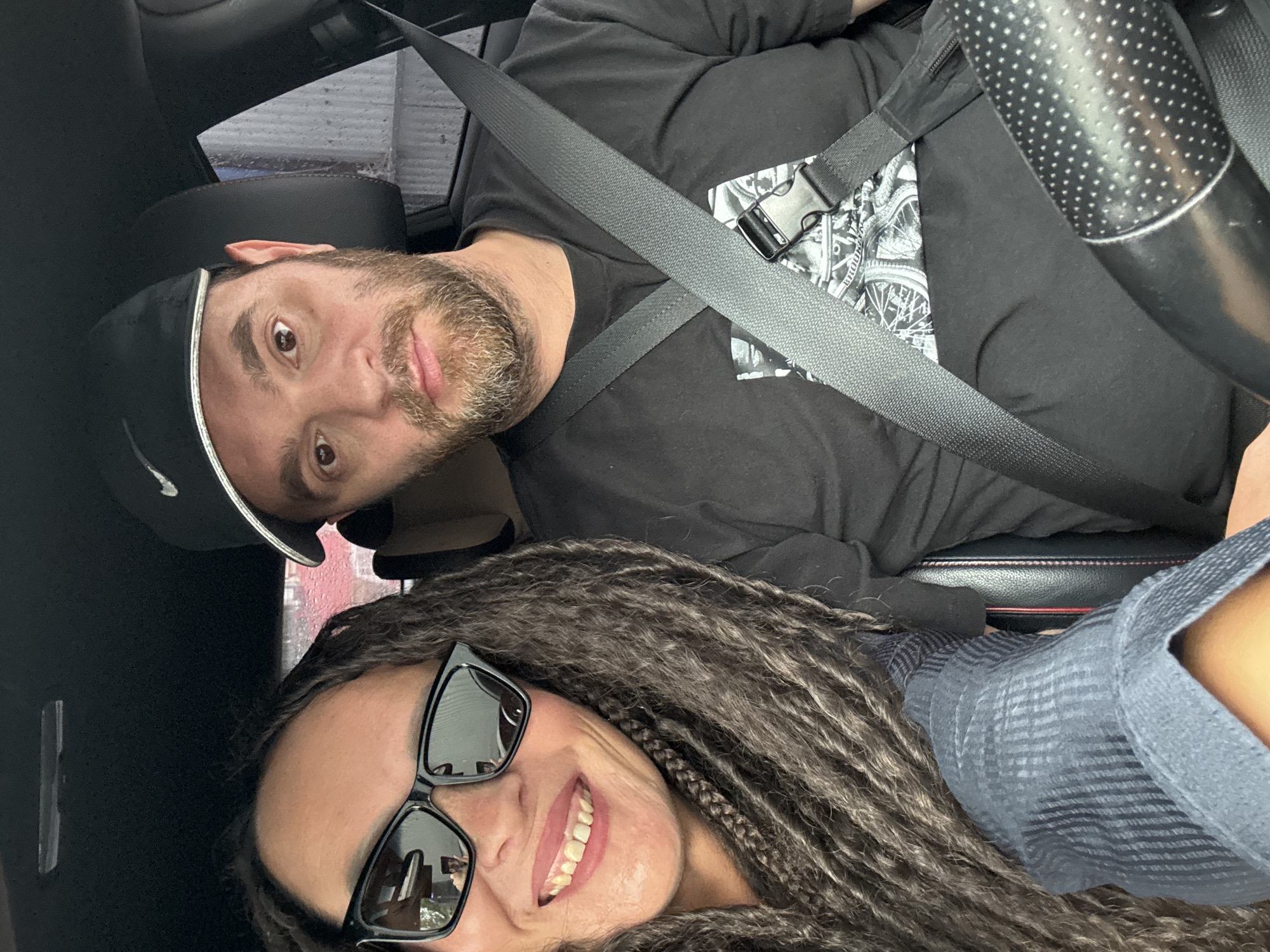 The image shows a selfie taken inside a car, featuring a man and a woman, oriented sideways (rotated 90 degrees counter-clockwise). The setting is likely Chaikovsky, Russia, within a vehicle during what appears to be daytime.
In the foreground and closer to the bottom of the frame, a woman is seen smiling broadly. She has long, dark dreadlocks or braids that frame her face and is wearing black sunglasses, which reflect parts of the car interior and exterior light. Her top is a light blue or gray patterned shirt.
Above and behind her, a man with a beard and a black baseball cap looks directly at the camera with a neutral to slightly amused expression. He is wearing a black t-shirt with a graphic design that includes bicycle wheels and the visible text "BROOKLYN". Both individuals are secured with black seatbelts, with the man's seatbelt clearly visible across his chest and shoulder, featuring a black plastic buckle.
A portion of the car's interior, including black seats and a textured steering wheel with small white dots, is visible. Through the car window in the background, a hint of the outside world, possibly a blurred building or sky, can be seen, suggesting the vehicle is either parked or in motion. The overall mood conveyed by the woman's smile is positive and cheerful.