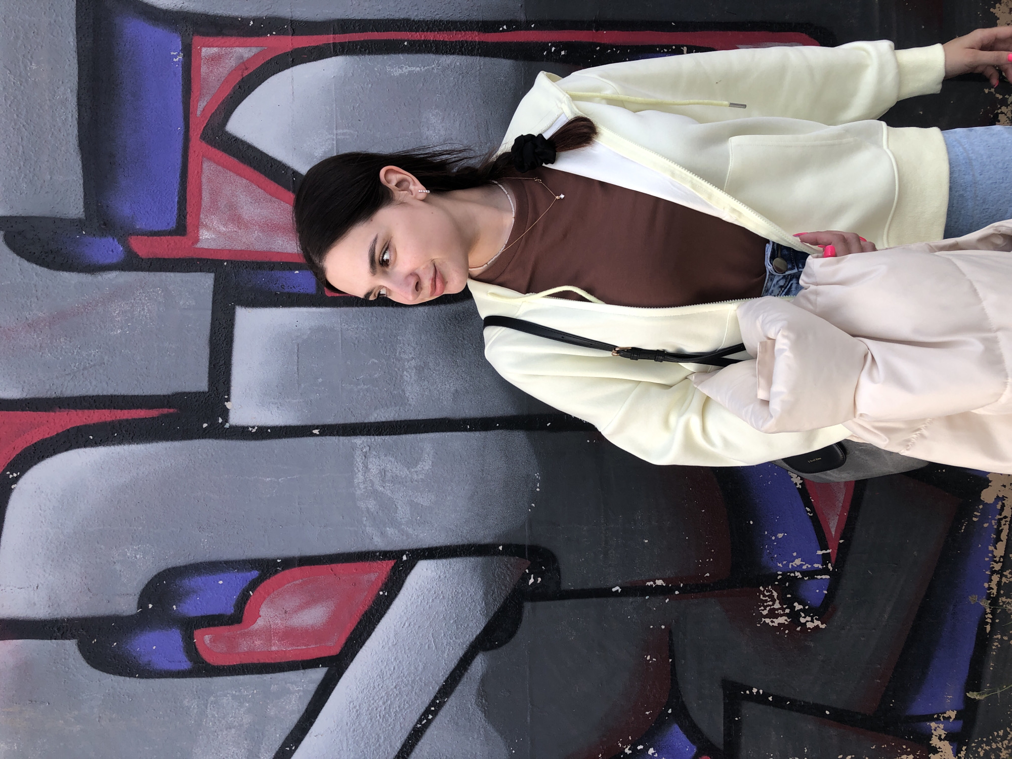 A young woman is pictured in a full-body shot, leaning against a graffiti-covered wall in an outdoor setting. The image is rotated 90 degrees counter-clockwise, showing the woman lying on her side relative to the frame. She has dark hair tied back with a black scrunchie, and wears small, sparkling earrings and a delicate necklace. Her gaze is directed slightly downwards and to her left, with a calm, somewhat contemplative expression.
She is dressed in a light yellow zip-up hoodie worn over a brown long-sleeved top and light blue pants or jeans. A light pink puffer jacket is draped over her right arm. Her fingernails are painted a bright pink. The background is a textured wall adorned with bold, abstract graffiti designs featuring large shapes outlined in black, filled with shades of gray, red, and purple. The lighting suggests it is daytime. This scene is located in Chaikovsky, Russia. There is no discernible text visible in the graffiti or on her clothing.