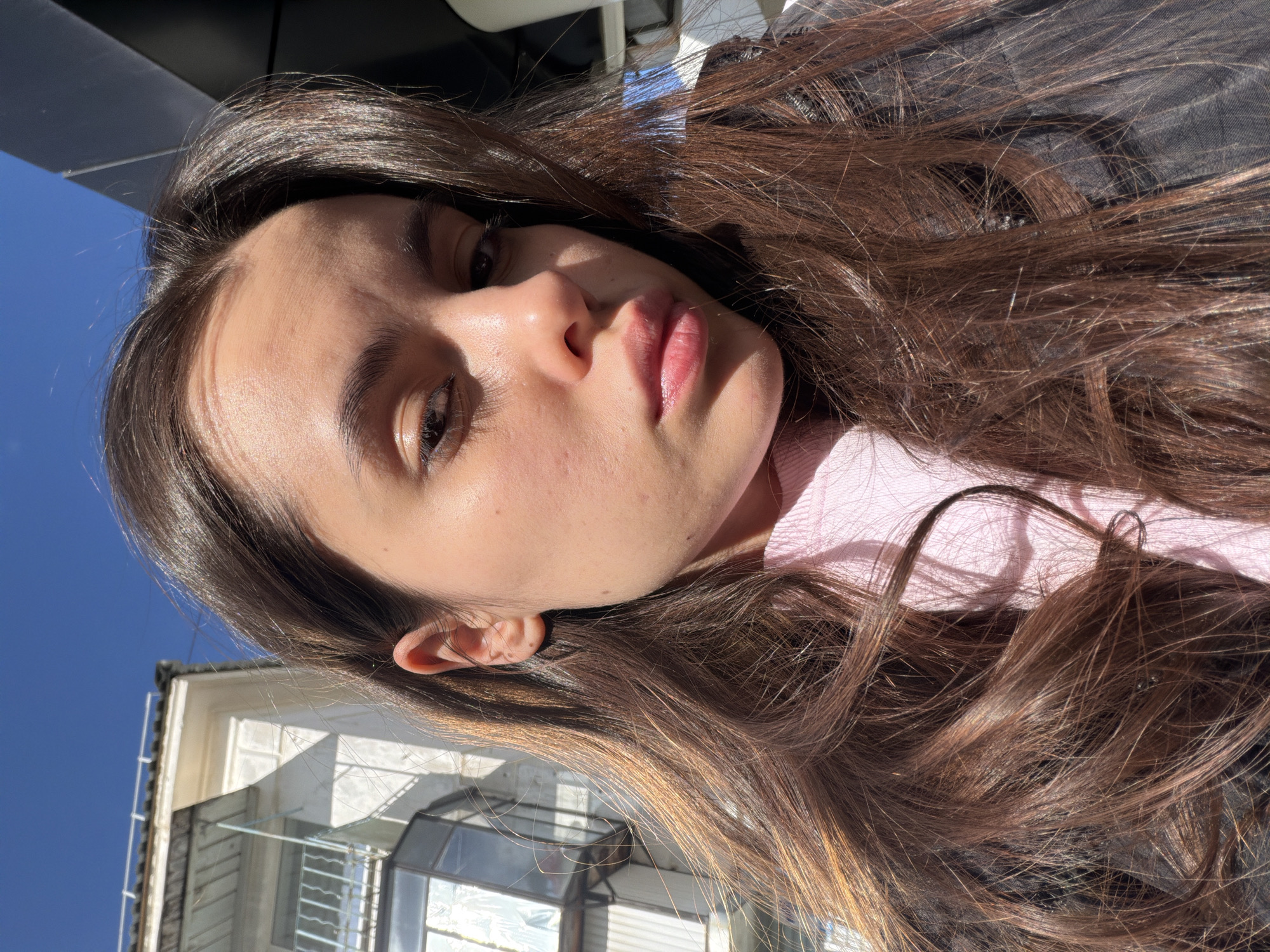 A young woman with long brown hair, wearing a pink top and a black jacket, poses for a selfie. Behind her, a building with white walls and window panes is visible against a clear blue sky. The location is Chaikovsky, Russia.