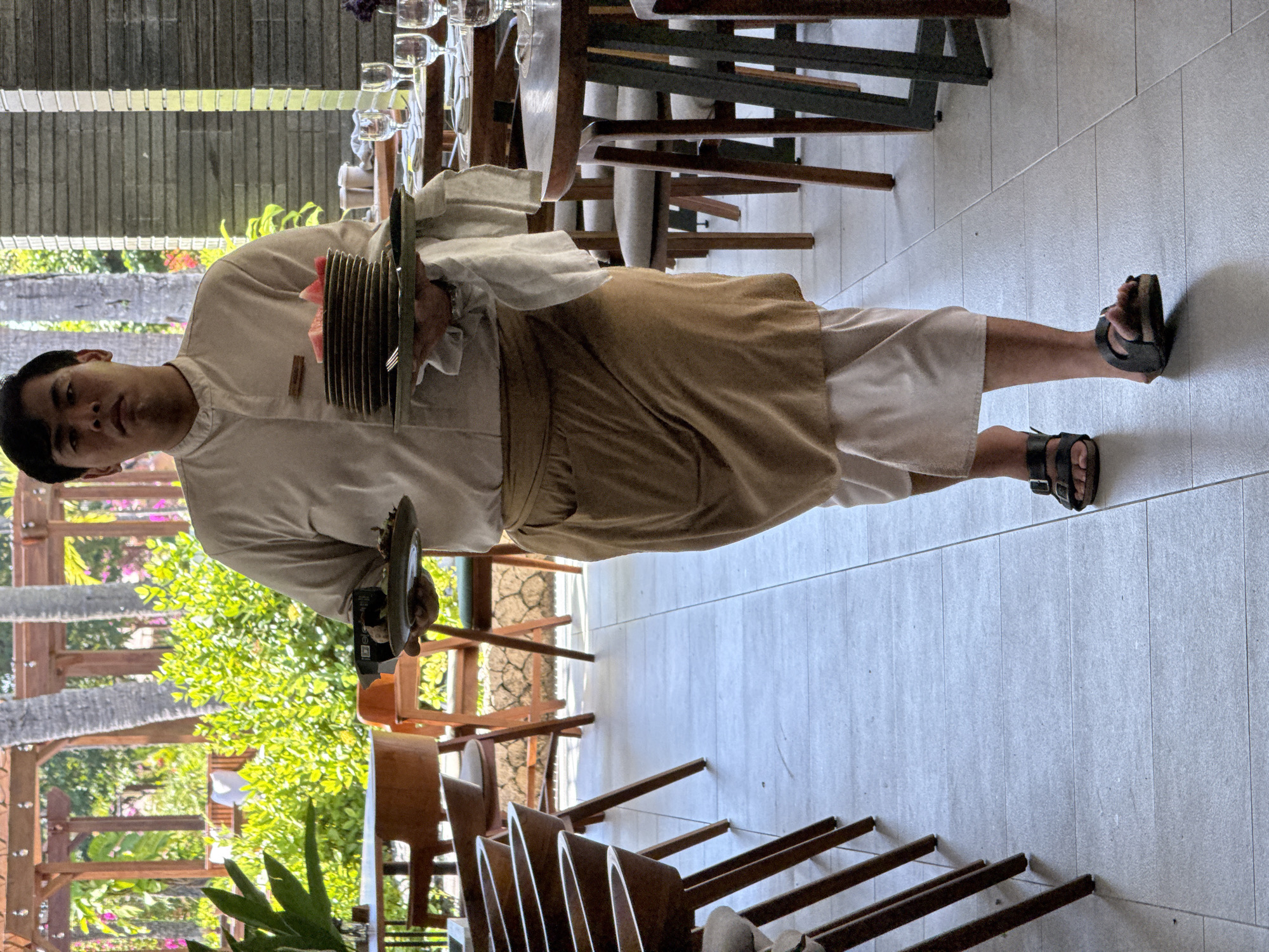 A full-body shot captures a staff member, likely a server, standing in a semi-outdoor dining area in Kuta Selatan, Indonesia. The individual, appearing to be male, has dark hair and a neutral expression, looking directly towards the viewer. They are dressed in a light-colored, long-sleeved uniform shirt, a brown waist-length apron or wrap skirt, light-colored trousers or capris, and dark sandals.
The person is actively engaged in carrying items; their left arm supports a stack of several brown plates, topped with a small pink or red decorative flower and possibly a piece of cutlery. Their right hand appears to be holding a tray or another flat item.
The setting suggests a restaurant or resort dining area. In the foreground and background, various wooden tables and chairs are visible, some neatly set with glasses, indicating preparation for service. The floor is covered with light-colored rectangular tiles. Beyond the dining area, lush green foliage and tropical plants are prominent, including the trunks of what appear to be palm trees. The bright natural light indicates it is daytime, with sunny weather. A wooden pergola or structural elements are also part of the outdoor environment. No discernible text is visible on the person's uniform or in the background.