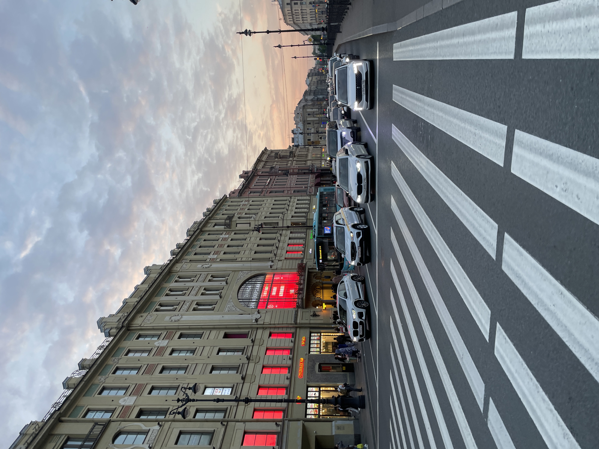 The image depicts a street scene in Saint Petersburg, Russia. A line of white cars is stopped in traffic on a street marked with a pedestrian crosswalk featuring bold white stripes. On the left, a multi-story building with a pale facade and architectural details dominates the frame. The building features large windows, some of which are illuminated with a bright red glow. The building's signage includes a visible "SKK" abbreviation, likely referring to the Petersburg Sports and Concert Complex. People are seen walking on the sidewalk in front of the building. The sky above is filled with clouds tinted with shades of pink and gray, suggesting either a sunrise or sunset. Street lamps are visible along the street.