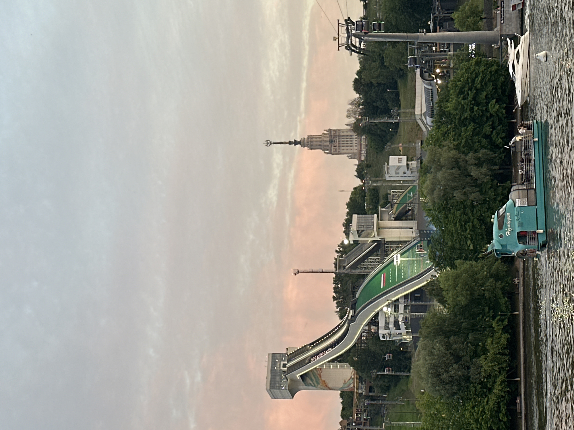 This is a wide shot of a body of water in Moscow, Russia, with a ski jump structure and a tall, Stalinist-era skyscraper in the background. The sky is a pale pink and gray, indicating it is either dawn or dusk. In the foreground, a turquoise boat with people on deck is moving across the water, and a white yacht is also visible in the middle ground. The ski jump is a prominent feature, with a steep ramp that transitions into a landing area. The skyscraper, with its distinctive spire, is a well-known landmark. Lush green trees surround the ski jump and the base of the skyscraper, suggesting a park or natural area. A cable car system is also visible, with gondolas moving across the frame. The overall impression is one of a scenic urban landscape at twilight.