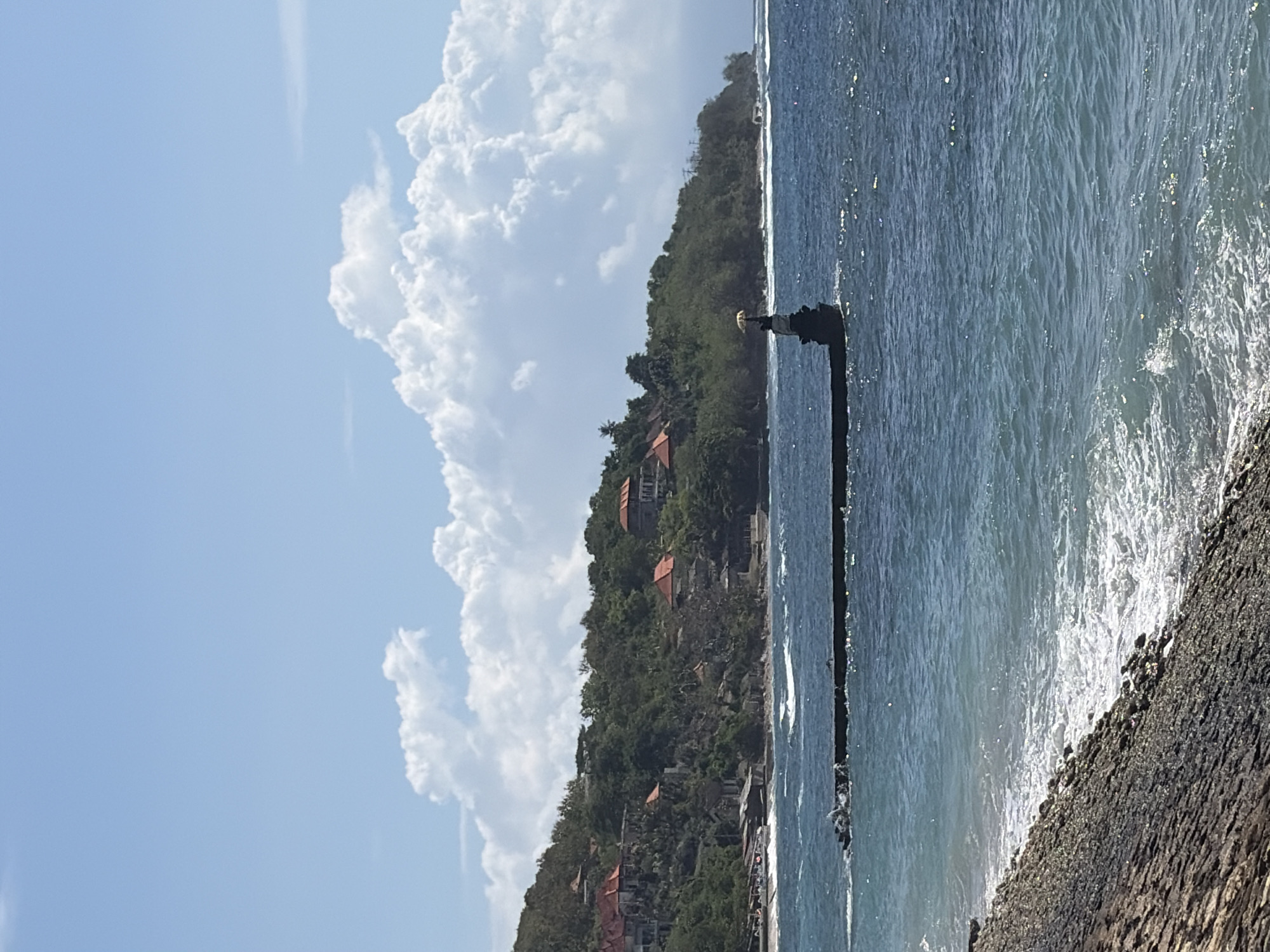 A view of the ocean and a tropical coastline in Nusa Penida, Indonesia. The foreground shows a rocky shoreline with small waves lapping at it. In the middle ground, a long, dark pier juts out into the blue water. At the end of the pier, a small, dark statue or monument stands, topped with a yellow umbrella. Beyond the pier, the coastline is lined with lush green trees and vegetation, with several buildings featuring red-tiled roofs nestled into the hillside. The sky is a clear blue with large, fluffy white clouds. The water is a deep blue, with whitecaps indicating a moderate choppiness. The scene is bathed in bright sunlight.