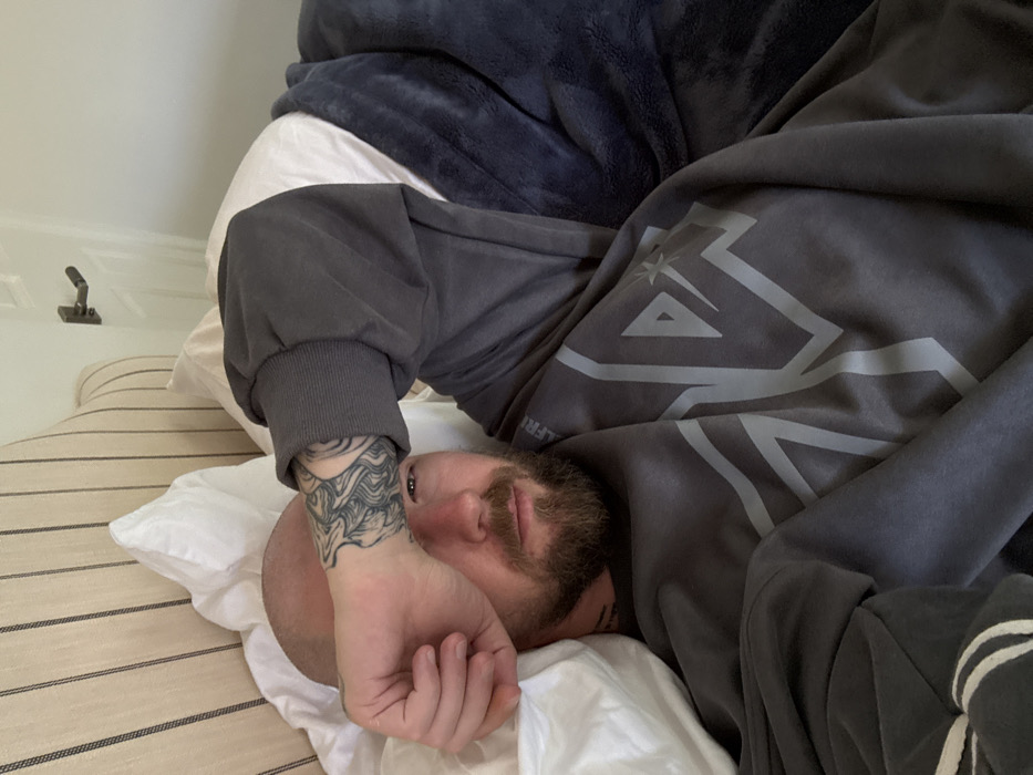 The scene is an indoor bedroom, located in a residence in Los Angeles, United States of America. An adult male with a bald head and a full beard is lying in a bed, looking directly at the camera. He is wearing a dark grey long-sleeved sweatshirt with a lighter grey abstract graphic design on the chest. His left arm is raised, with his hand covering the upper part of his face, including his left eye and forehead, possibly shielding from light. A prominent black and grey tattoo with swirling, organic patterns is visible on his left forearm and wrist. He is resting on white pillows. A striped, light-colored upholstered headboard is visible behind him. To his right, a dark blue, plush blanket or throw covers part of his body. In the upper right background, a white door with a dark-colored lever door handle is visible.