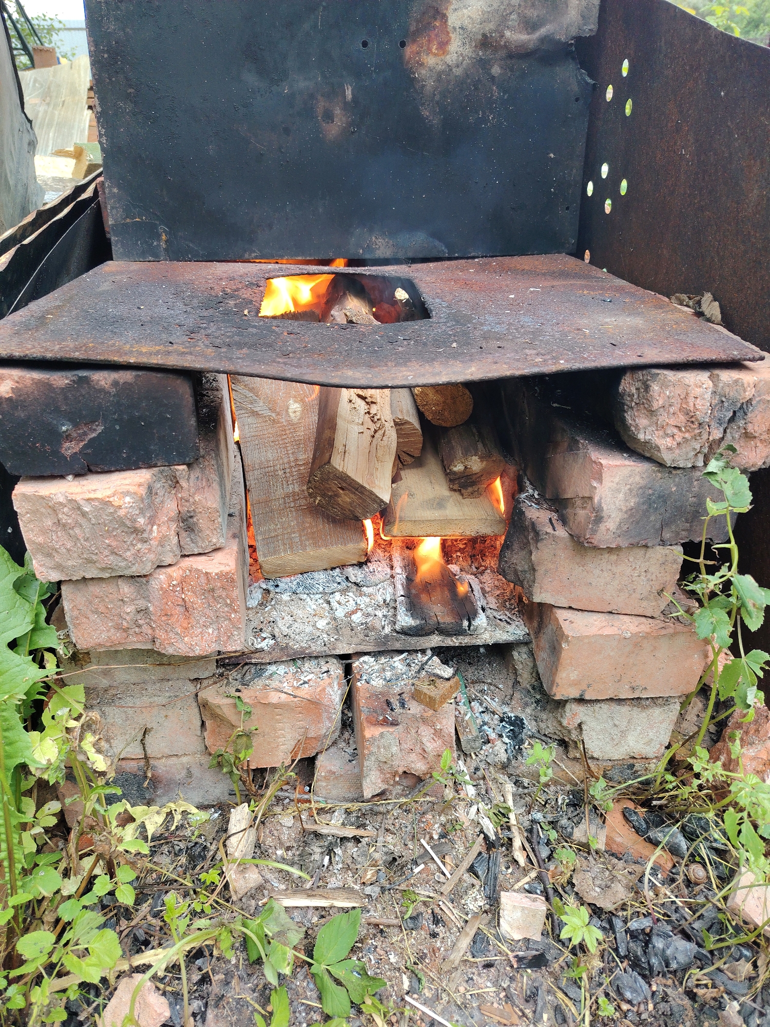 The image shows a makeshift brick oven with a fire burning inside. The oven is constructed from red bricks stacked to form a combustion chamber, with a flat piece of rusted metal serving as the top and a cooking surface. Inside the chamber, pieces of wood are burning, with flames and glowing embers visible through an opening in the metal top. The area around the oven is covered in dirt, ash, and sparse green vegetation, suggesting an outdoor setting.
The scene appears to be an informal cooking setup, possibly in a rural or backyard environment. The presence of ash and burnt wood indicates that the oven has been used recently or is currently in operation. The lighting suggests it is daytime, but there are no strong shadows to indicate a specific time. The overall impression is one of rustic, functional resourcefulness. No people are visible in the image, nor is any text. The location context provided is Мшинская, Russia.