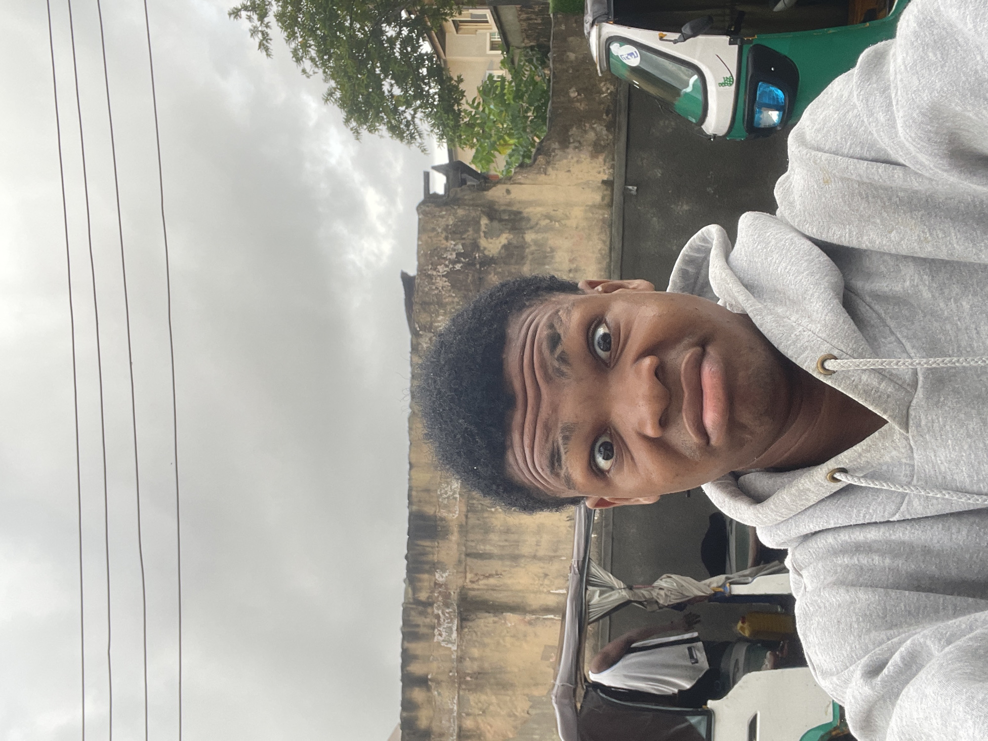 The image depicts an outdoor selfie taken in Ado, Nigeria, featuring a young Black male from the chest up. He wears a grey hooded sweatshirt with visible white drawstrings and metal grommets. His facial expression is exaggerated, characterized by wide-open eyes, raised and furrowed eyebrows, and tightly pursed lips. His hair is short, dark, and curly.
The background shows an urban or semi-urban environment under an overcast, grey sky, crossed by four dark utility lines. A weathered, light-brown concrete wall with visible discoloration and texture spans horizontally across the mid-ground. To the viewer's left, a portion of a white and green tricycle (auto-rickshaw) is visible, with a second male figure in a white t-shirt partially obscured within or near it. A yellow container rests on the ground near this vehicle. To the viewer's right, the front portion of another green and white tricycle is discernible, featuring blue-tinted headlights. Behind this, green foliage and the light-colored facade of a building are visible further in the distance. The ambient lighting is diffused, consistent with heavy cloud cover.