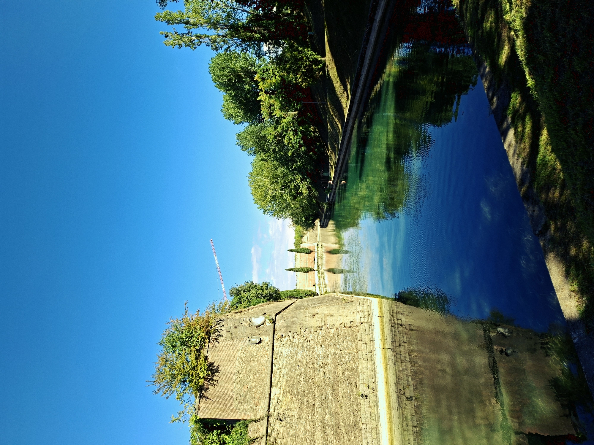 The image depicts a view of the moats surrounding the historic fortress in Peschiera del Garda, Italy. The clear blue sky dominates the upper portion of the frame, with a faint contrail from an airplane visible. A prominent brick wall of the fortress, overgrown with vegetation, runs along the left side of the image, its reflection mirrored in the calm, emerald green water of the moat. The moat is bordered by grassy banks and a concrete walkway on the right, with dense green trees lining the opposite bank of the moat, casting shadows on the grass. Several cypress-like trees stand along the fortress walls in the middle distance. The water's surface is calm, with ripples indicating a gentle current. A distant, red and white construction crane is visible above the fortress walls on the right side of the image. The overall scene conveys a sense of historical grandeur and natural beauty under a bright, sunny sky.