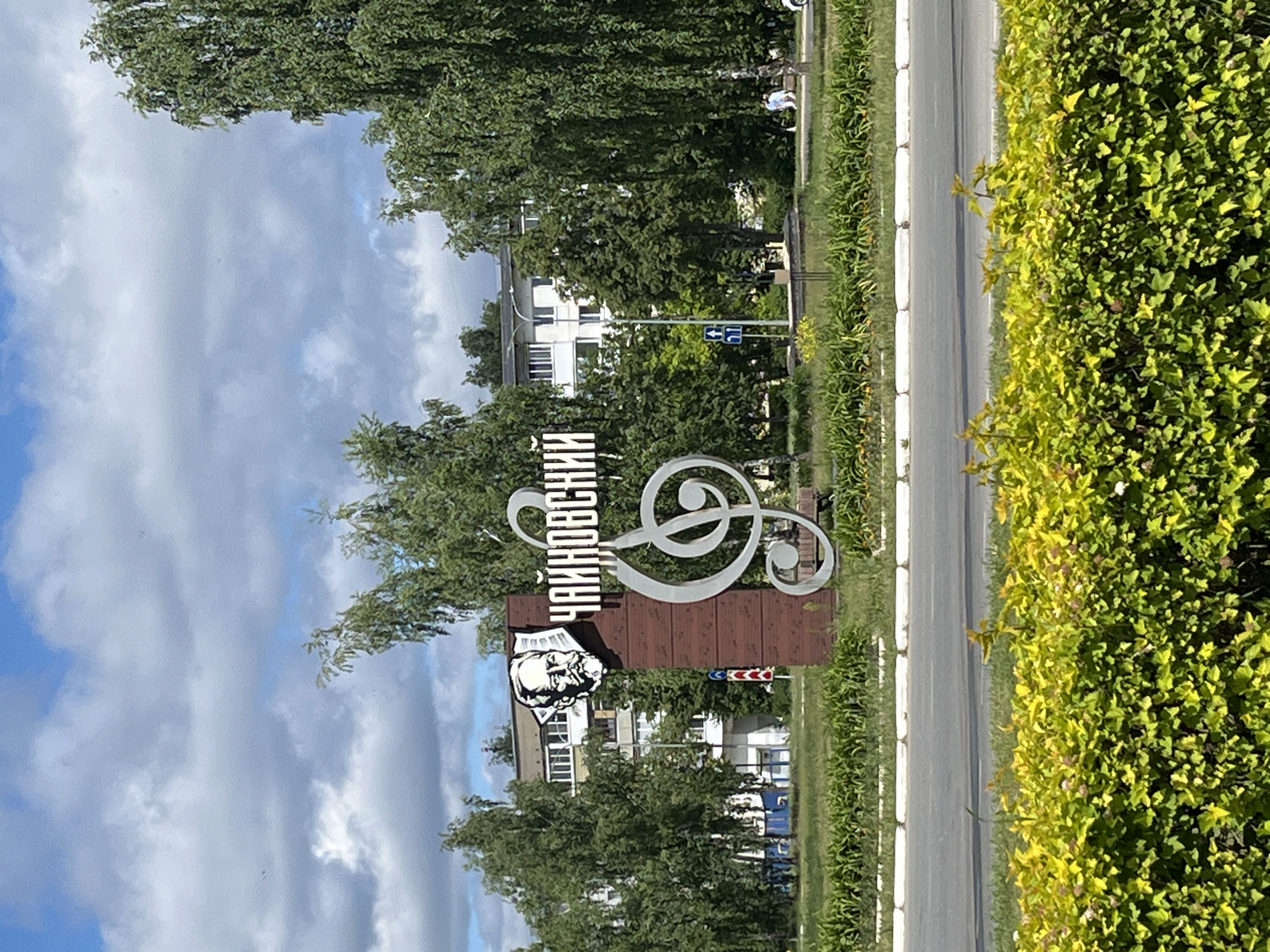 A vertical, eye-level, medium shot shows a large monument sign in the city of Chaikovsky, Russia. The sign is brown with a portrait of Pyotr Ilyich Tchaikovsky and the Cyrillic inscription "Чайковский" in white lettering. A large treble clef made of polished metal is attached to the sign. In the background, there are several large trees, some apartment buildings, a street, and a green lawn. A few people are visible walking in the distance. The sky is blue with some scattered clouds. In the foreground, there is a hedge of green and yellow leaves.