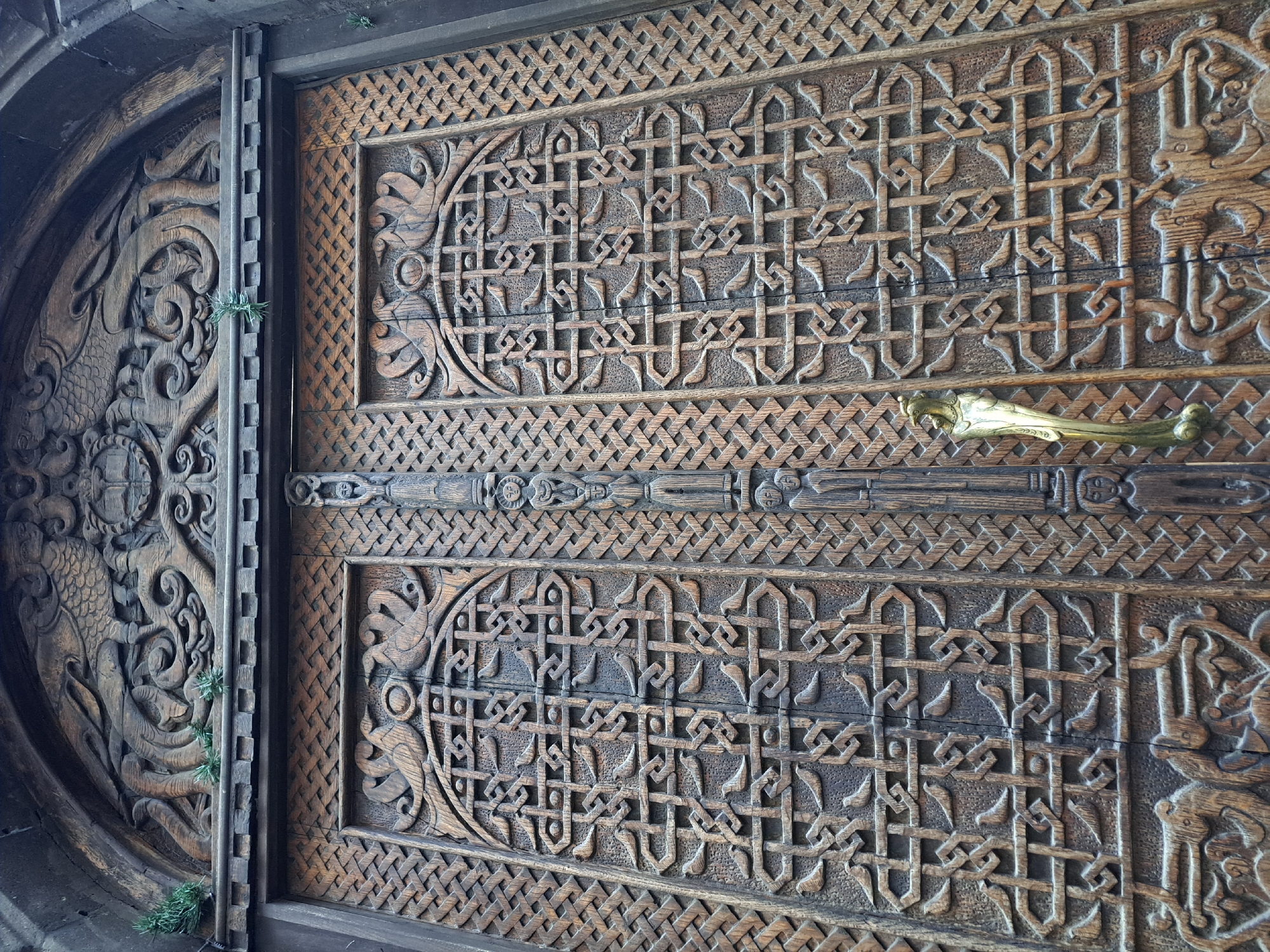 A close-up photograph reveals a heavily carved wooden door with intricate designs. The door is composed of multiple panels. The left panel features a rounded edge with a scene of carved sea serpents or dragons. The central and right panels contain geometric patterns and arabesque forms. A thick, carved horizontal bar runs across the center of the central panel, with a decorative metal handle attached. Small tufts of green vegetation are visible between the edge of the left and central panels. The door is possibly located in Yerevan, Armenia.