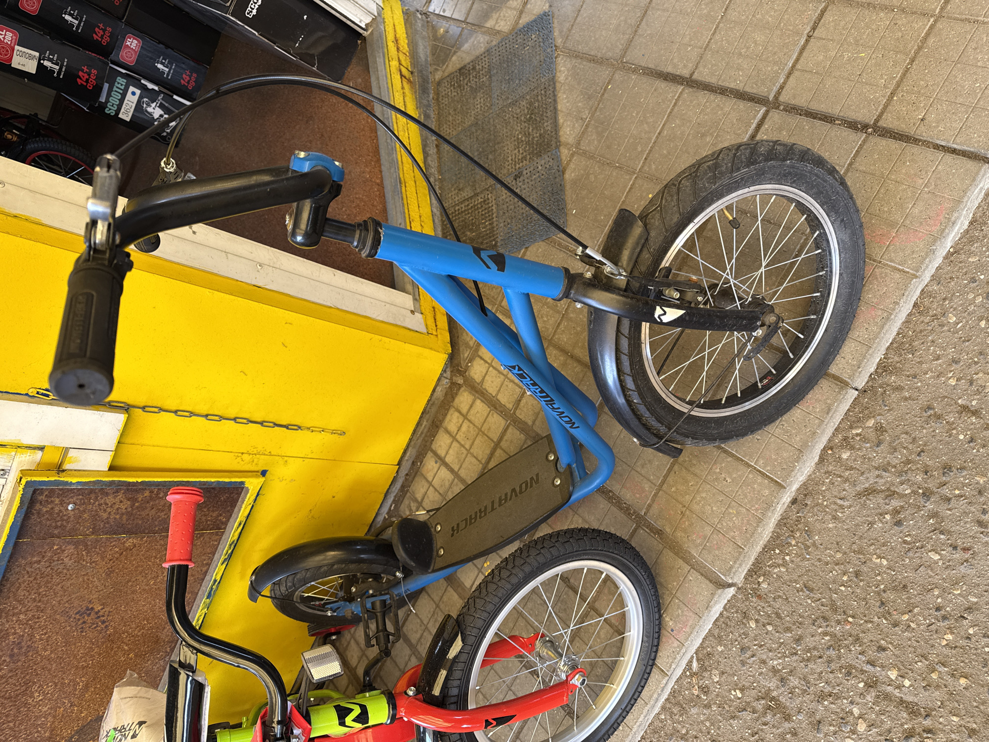 The image shows two Novatrack scooters, one blue and one red, parked on a concrete tiled surface near a yellow wall. The blue scooter is positioned vertically with its front wheel on the right, angled towards the viewer, and its handlebars near the top left corner. The red scooter is on the left side, slightly obscured, with its handlebars featuring red grips. Behind the yellow wall, stacked boxes labeled "SCOOTER," "XL 200," and age ranges are visible. The scooters are stationary, and the environment appears to be an urban setting.