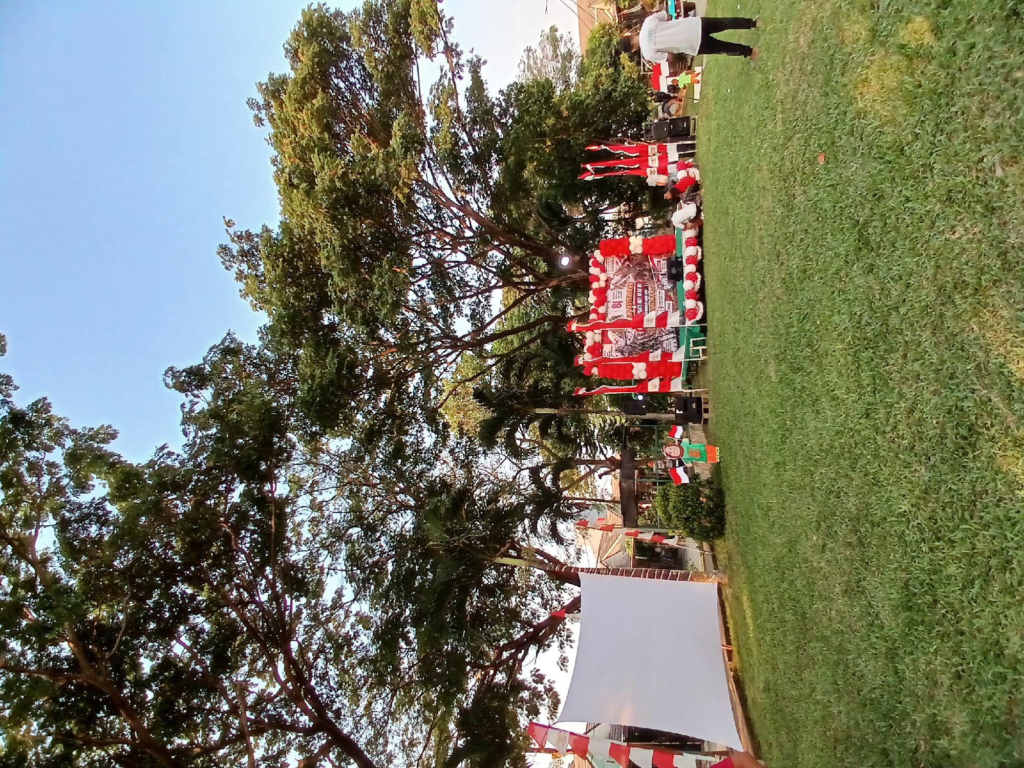 In the city of Kebalen, Indonesia, an outdoor stage has been set up on a grassy field under the shade of large trees. The stage is decorated with red and white balloons and banners, and appears to be prepared for an event. In front of the stage, a large white screen is set up on a wooden frame, possibly for a projection. Several people are gathered on and around the stage, some are seated, and others are standing. A man in a white t-shirt and black pants stands facing away from the camera in the foreground on the right side of the image, observing the setup. The overall atmosphere suggests a community event or celebration is about to take place.