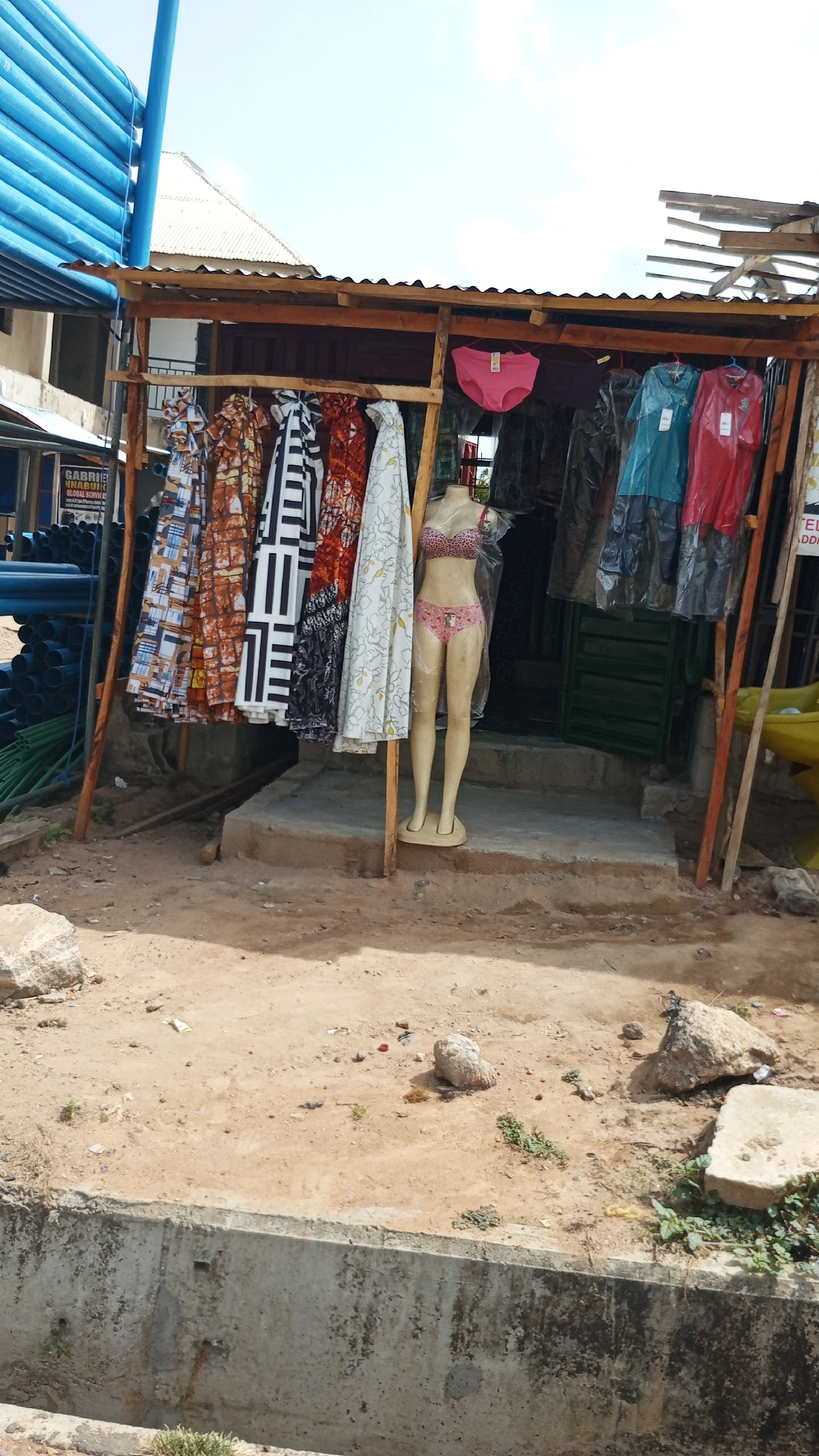 Outside a ramshackle stall in Zango-ndaji, Nigeria, a mannequin dressed in a pink bikini stands at the stall entrance. Garments on hangers are displayed along the front of the stall. Clothing in plastic bags hangs further inside. The stall appears to be constructed of wood and corrugated metal. The foreground is a dirt surface with debris and rocks. A concrete drainage ditch runs along the bottom edge of the frame.