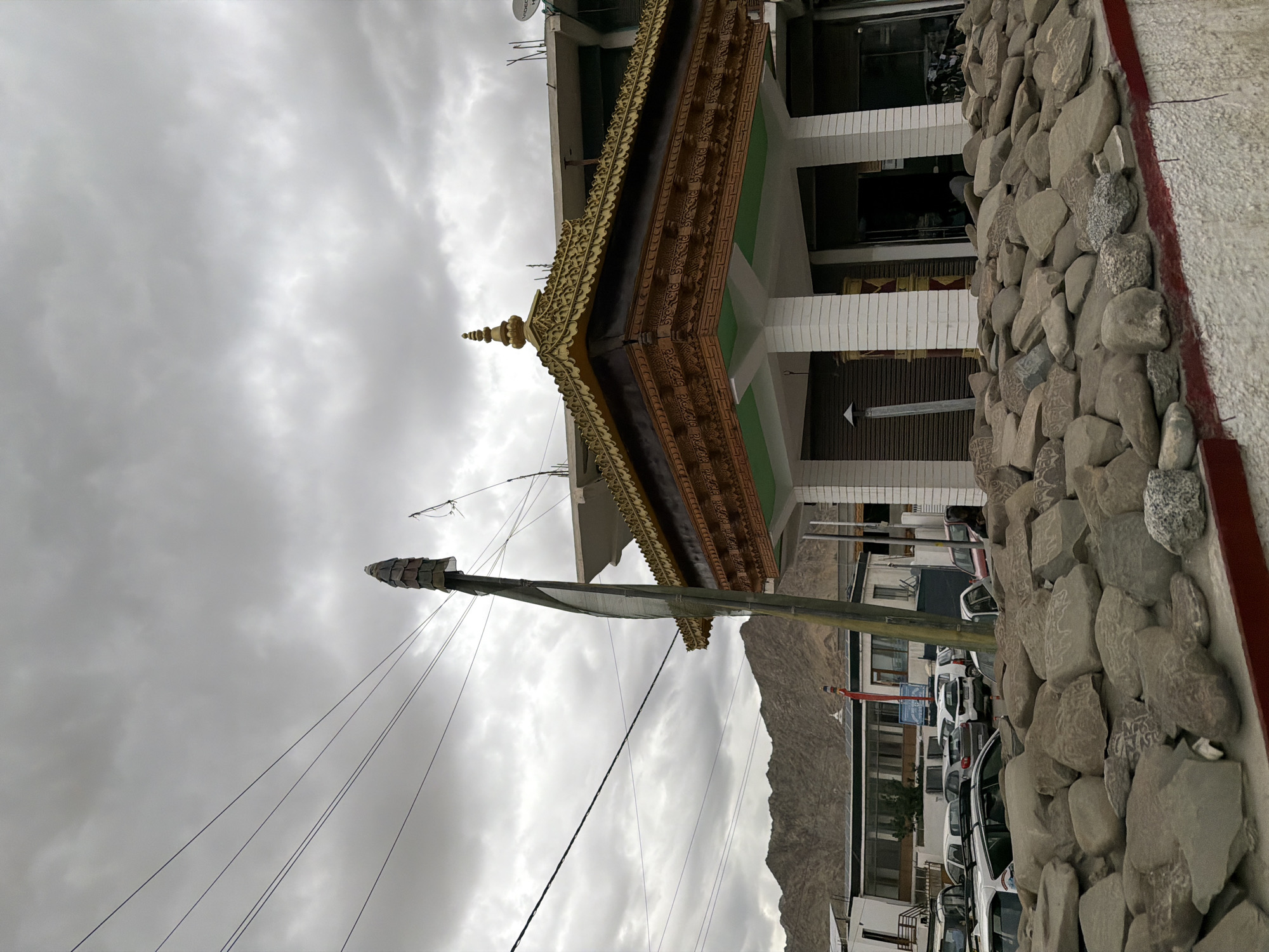In Leh, India, a cloudy sky looms over a street scene. In the foreground, a low wall is topped with a pile of large, flat stones, many of which appear to be carved with Tibetan Buddhist inscriptions. Behind the wall, a modern building with a golden, intricately carved roof supported by white columns stands to the right. To the left of this structure, a tall wooden pole with prayer flags is visible, along with power lines stretching across the sky. Further left, a row of parked cars is lined up in front of a building complex. In the background, a range of rugged, barren mountains rises under the overcast sky. The overall impression is one of a busy street in a Himalayan town, with a mix of modern infrastructure and traditional religious elements.