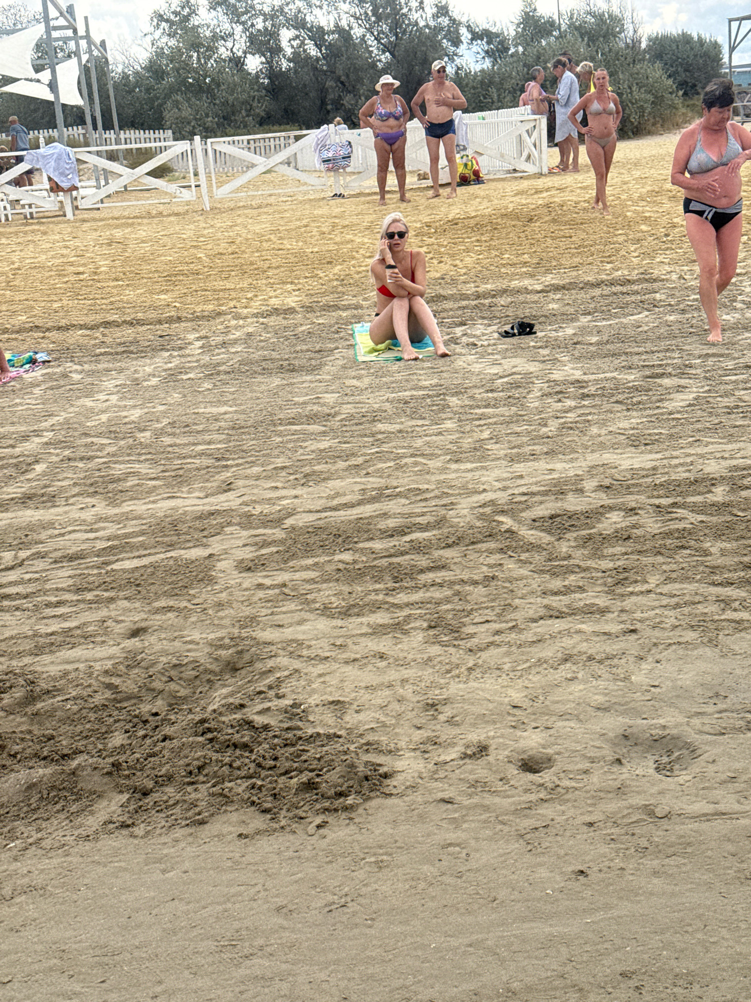 The image depicts a sandy beach with several people present. In the foreground, a woman in a red bikini is sitting on a towel, holding a phone to her ear. To her right, a woman in a black and white bikini walks away from the camera. In the background, a group of people are standing near a white fence. Among them are a woman in a patterned bikini and hat, a man in swim trunks, a woman in a light blue cover-up, and other individuals. The environment appears to be a public beach with some recreational structures visible, such as a white tent-like canopy and a white fence. The ground is covered in sand, with some textured areas indicating it may have been raked or disturbed. The sky is overcast. The scene is likely located in Anapa, Russia, given the presence of a beach with a sandy shore and recreational amenities commonly found in such locations.