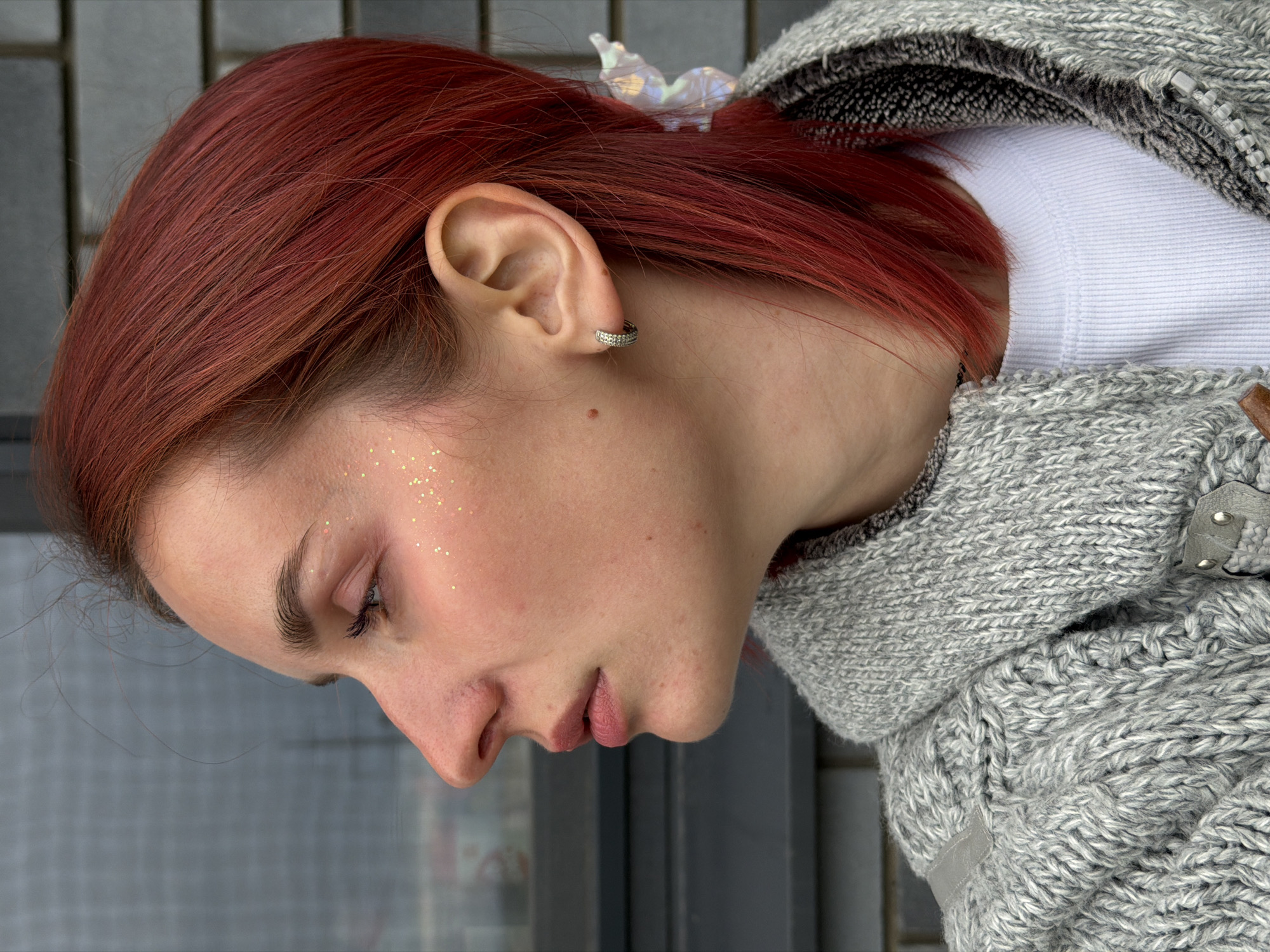 The image shows a close-up of a woman with red hair, wearing a gray knitted sweater over a white shirt. She is looking down with her eyes partially closed. There is gold glitter on her cheek. She has an earring in her left ear. The background appears to be a blurred window and possibly a wall with some texture. While the prompt asked to identify if the image was taken in Chaikovsky, Russia, this cannot be confirmed from the visual information present.