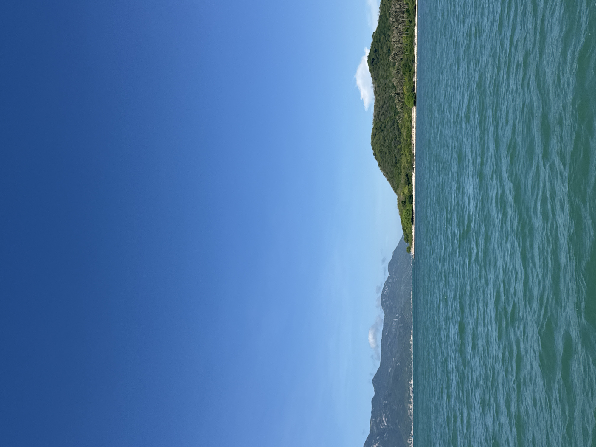 The media file depicts a seascape with a clear blue sky and a body of water. The scene is dominated by a vast expanse of blue sky occupying the left half of the frame, with a single, wispy cloud visible near the horizon.
On the right side of the image, a lush, green mountainside slopes down towards the water. The vegetation appears dense and tropical. At the base of the mountain, a narrow strip of light-colored land, likely a beach or shoreline, is visible. The water itself is a deep turquoise color, with gentle waves creating a textured surface. The water meets the shore in a calm manner.
The image is oriented vertically, suggesting it was taken from a boat or a viewpoint elevated above the water. There are no people or man-made objects visible in the scene, emphasizing the natural landscape. The time of day appears to be daytime, with ample sunlight creating a bright and clear atmosphere. The weather seems fair and pleasant. The overall impression is one of tranquility and natural beauty. No text is visible in the image.