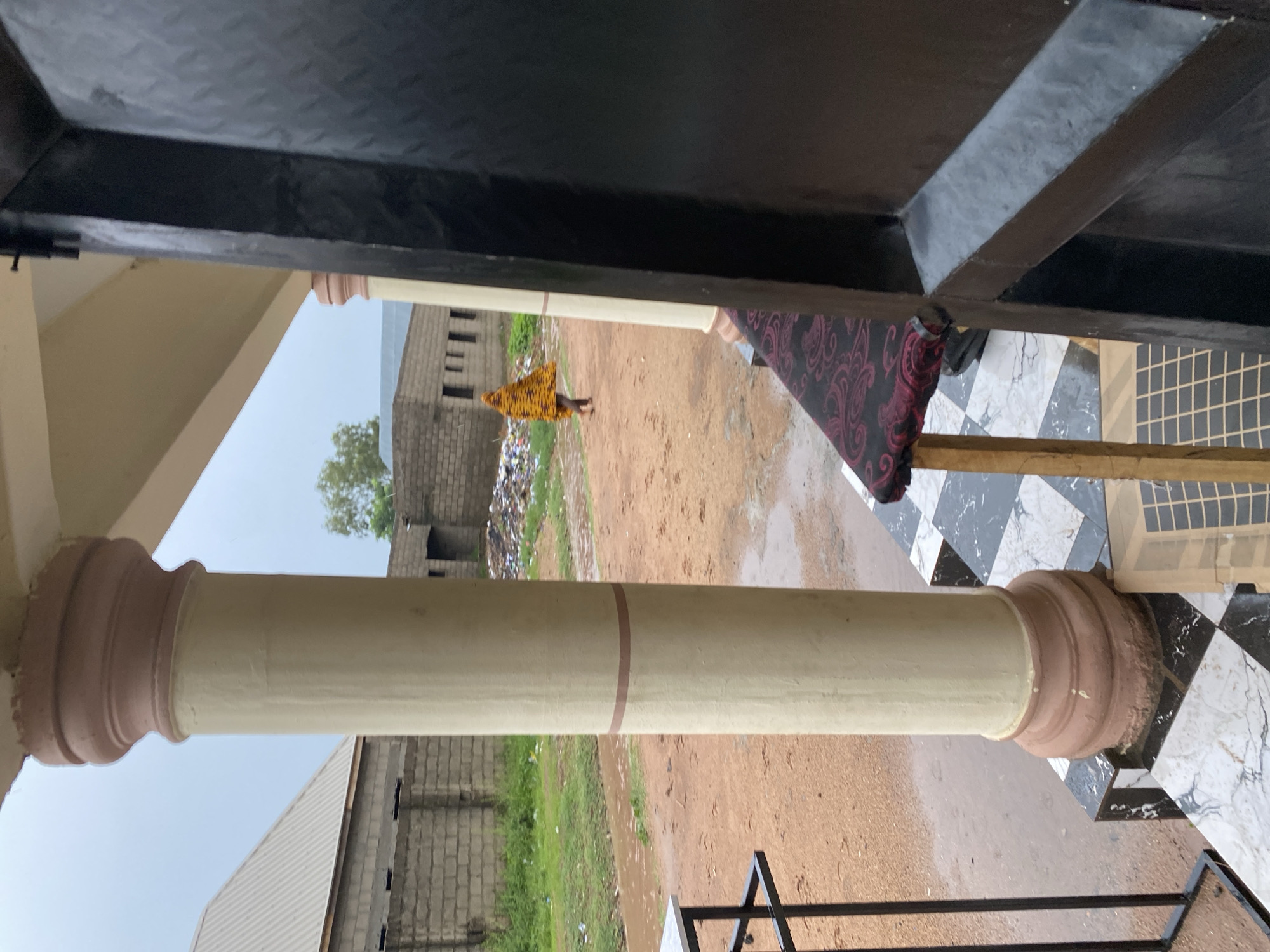 The image captures an outdoor scene in Jalingo, Nigeria, likely a residential or commercial area on a cloudy day. In the foreground, a large, cream-colored cylindrical pillar with a reddish-brown base stands to the left, partially obscuring a dark, textured vertical surface on the right, possibly a door or wall. The ground in front of the pillar is paved with black and white checkered tiles. Beyond the pillar, a dirt path is visible, with puddles indicating recent rain. In the mid-ground, a person is walking away from the camera, wearing a vibrant yellow and orange patterned garment that drapes over their head and body. Behind the person, there is a pile of debris or trash. Further in the background, there are buildings constructed from grey bricks, one with a corrugated metal roof. Green grass and some scattered litter are present in the middle distance. The overall atmosphere is somewhat unkempt, suggesting an area in need of development or cleanup.