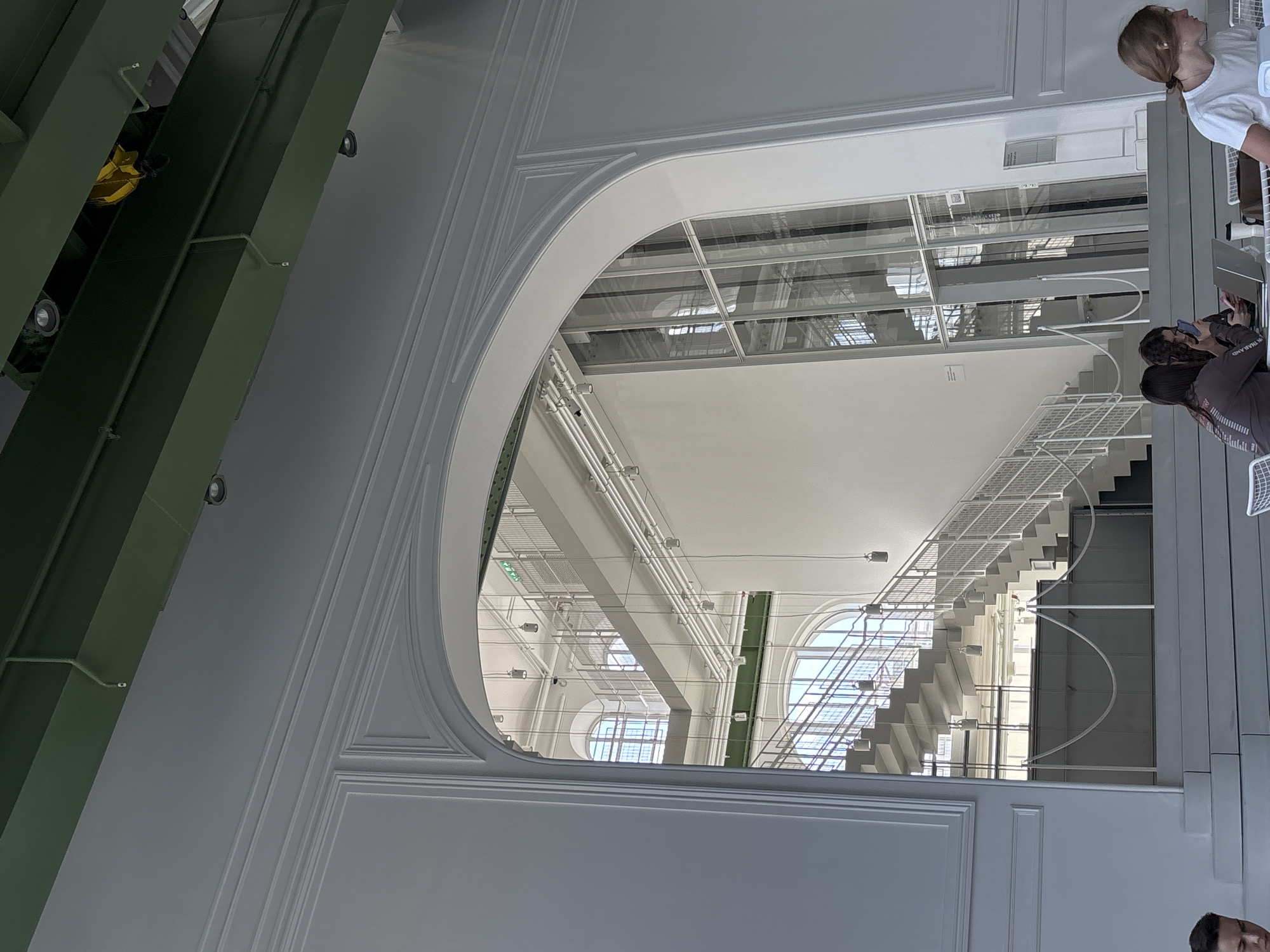 The image shows an interior view in Moscow, Russia, seen through a large arched opening within a white wall. On the left, a green structure extends diagonally from the top. The view through the arch reveals a large, bright space with a white ceiling and multiple levels of architecture. A white, metal staircase winds upward, with additional arches and windows visible in the background. Several people are positioned on the right side of the frame, near the viewer's location. One person is standing and wearing a white shirt, while others appear to be looking at phones or interacting near the staircase.