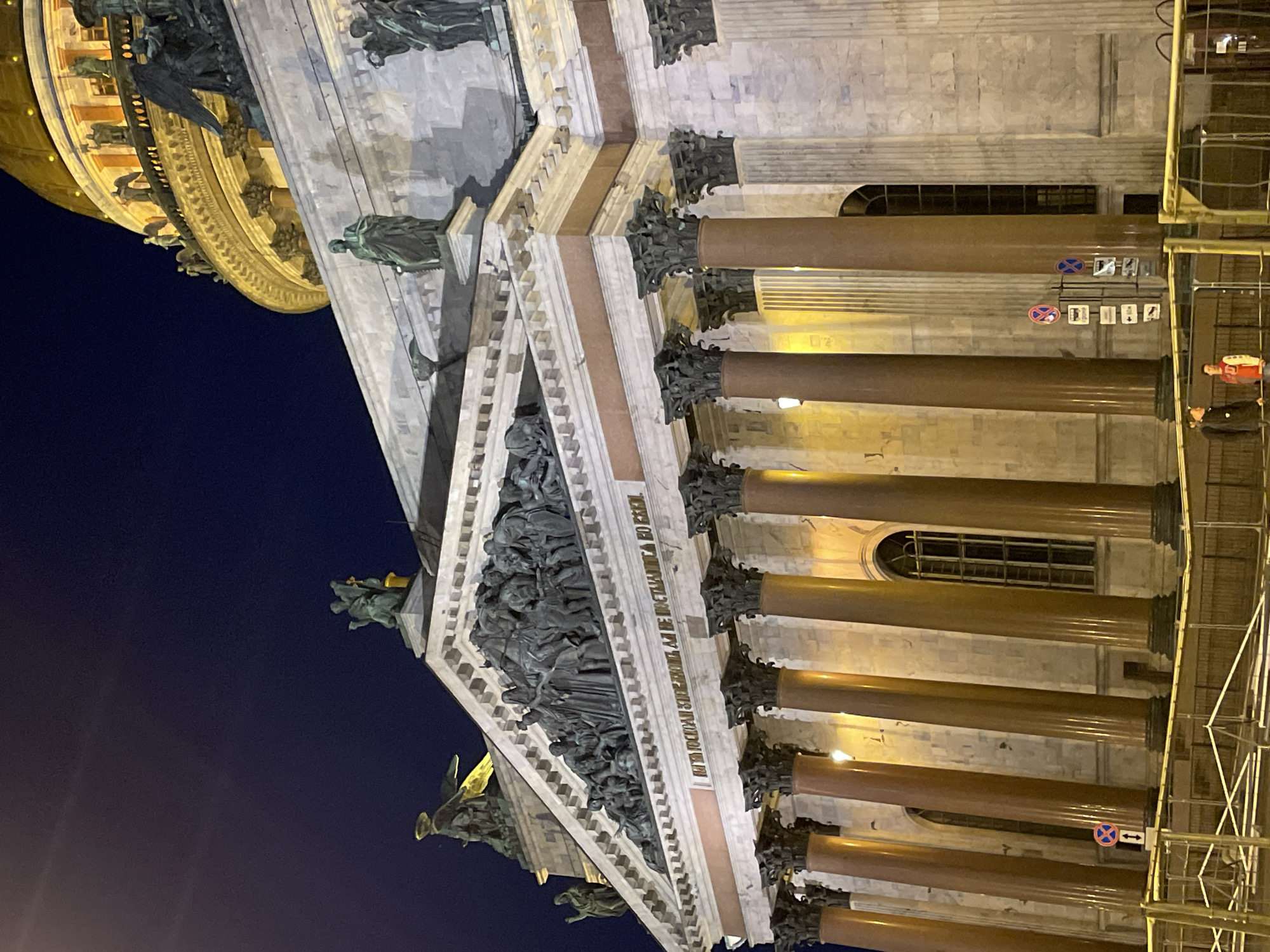 The image captures a detailed view of a grand classical building, identified as St. Isaac's Cathedral in Saint Petersburg, Russia, at night. The impressive facade is illuminated by artificial lights, casting a warm glow on the stone and bringing out the architectural details.
The primary subject is the building's portico, featuring massive, polished reddish-brown granite columns with ornate, dark capitals. Above the columns, a frieze with visible Cyrillic text reading "МНѢ ПОСТОИМЪ ... СЕЙ СОБОРЪ" (To me, let stand this cathedral) leads to a triangular pediment. The pediment is densely filled with dark, sculptural figures, depicting a complex scene. More sculptures adorn the roofline and the edge of a massive, partially visible golden dome or rotunda in the background.
A dark night sky forms the backdrop, contrasting sharply with the brightly lit structure. Arched windows are visible between some columns. On the right side of the image, a metal scaffolding structure is erected next to the building, partially obscuring it. Two individuals, one wearing a red top and another in a dark jacket, are visible standing near the scaffolding. Several circular street signs are present, including a "No Stopping" sign (blue circle with red diagonal line), a "No Entry" sign (red circle with white bar), and a blue directional sign with a white arrow. The overall scene suggests a nighttime visit or viewing of this iconic landmark.